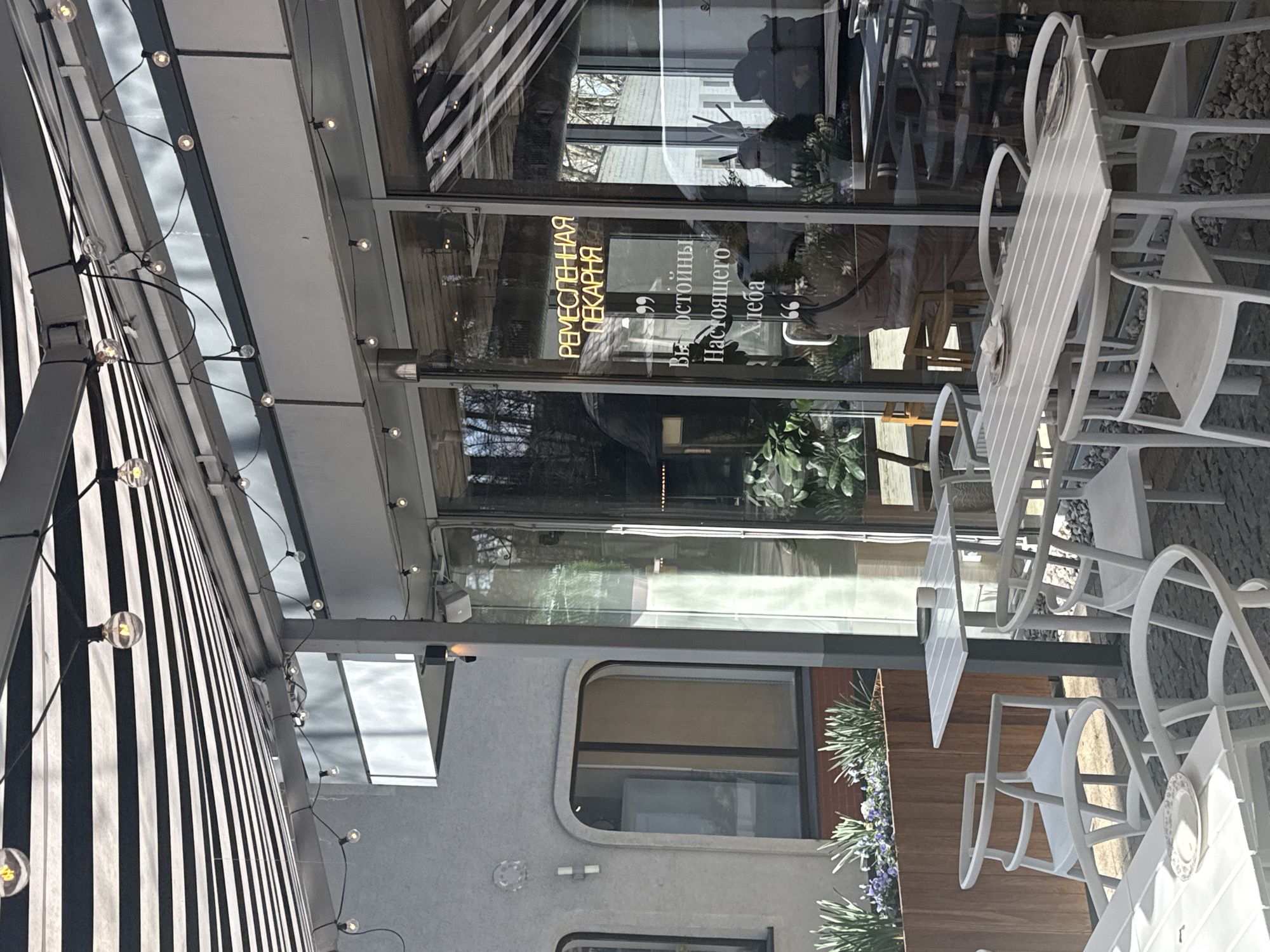 The image captures an outdoor seating area, likely a patio, in Svetlogorsk, Russia. The seating area is located next to a building with a glass facade. On the glass, there are signs in Russian. The text on the signs appears to say "Artisan Bakery" and "worthy of real bread." Adjacent to the facade is a striped awning with strings of lights, adding a decorative touch. The seating area includes white tables and chairs. Some tables have plates on them, suggesting they're set for customers. Planters with foliage add greenery to the space. The environment appears to be an urban setting, reflecting details of the city in the reflections of the windows.