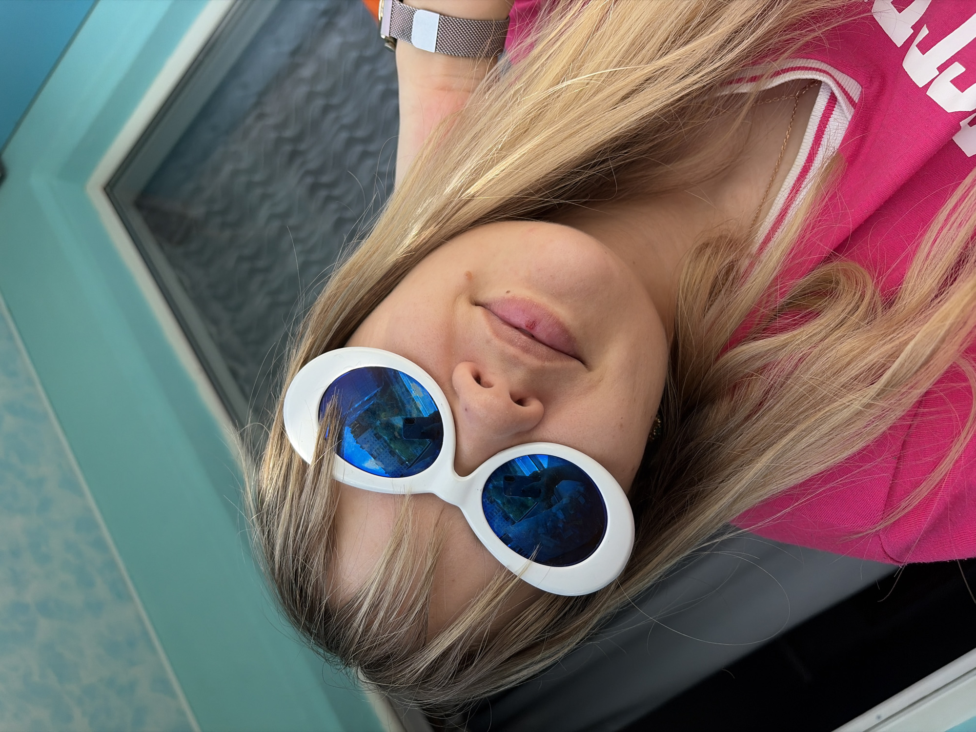 A fair-skinned woman with long blonde hair wears sunglasses with white frames. The sunglasses reflect what appears to be a blue, multi-story building. She is wearing a pink shirt and a metallic watch. The background shows part of a window frame and light blue wall or surface. Given that the location is Chaikovsky, Russia, the reflected building might potentially be one in the city, although without further visual cues, it's difficult to confirm this with certainty.