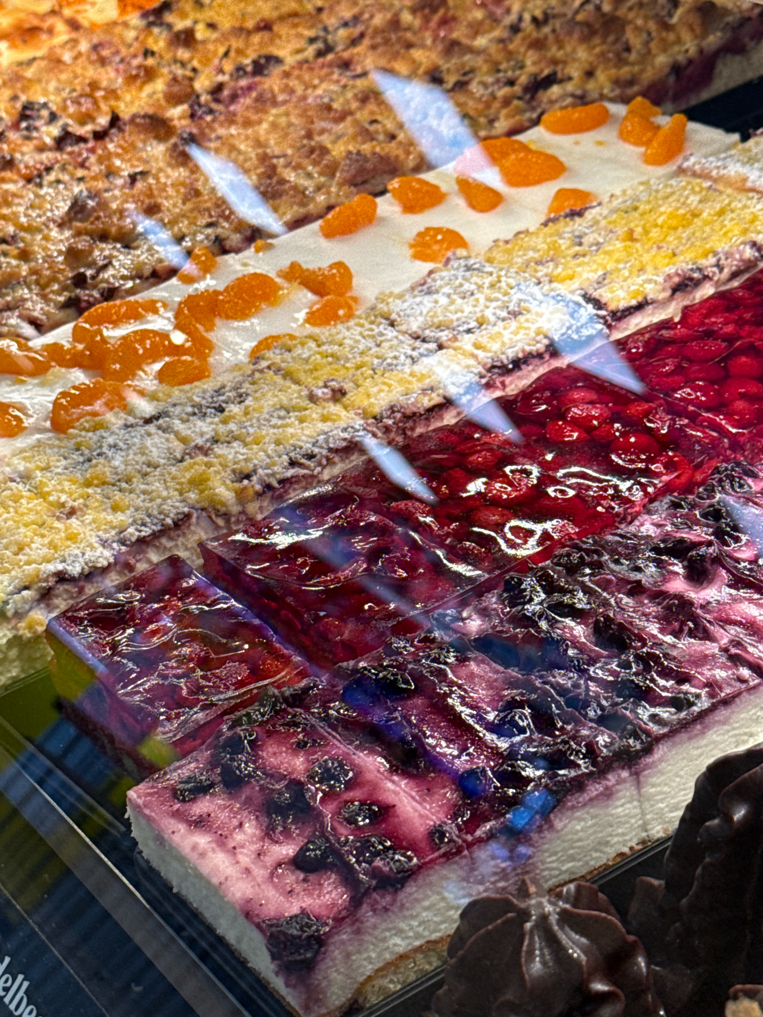The image shows a display case filled with various types of cakes and pastries. In the foreground, several slices of cheesecake topped with blueberries and a glossy purple glaze are visible. To the right of the cheesecake, a dark chocolate pastry with a decorative swirl is partially in view. Behind the cheesecake, there are more cake slices, including one with a bright red berry topping, possibly cherries or raspberries, and another with a yellow crumble topping and mandarin orange segments. A golden-brown cake with dark fruit pieces and a streusel topping is visible at the top of the image. The overall impression is a bakery display showcasing sweet treats. The location is a bakery in Herzberg, Germany.
