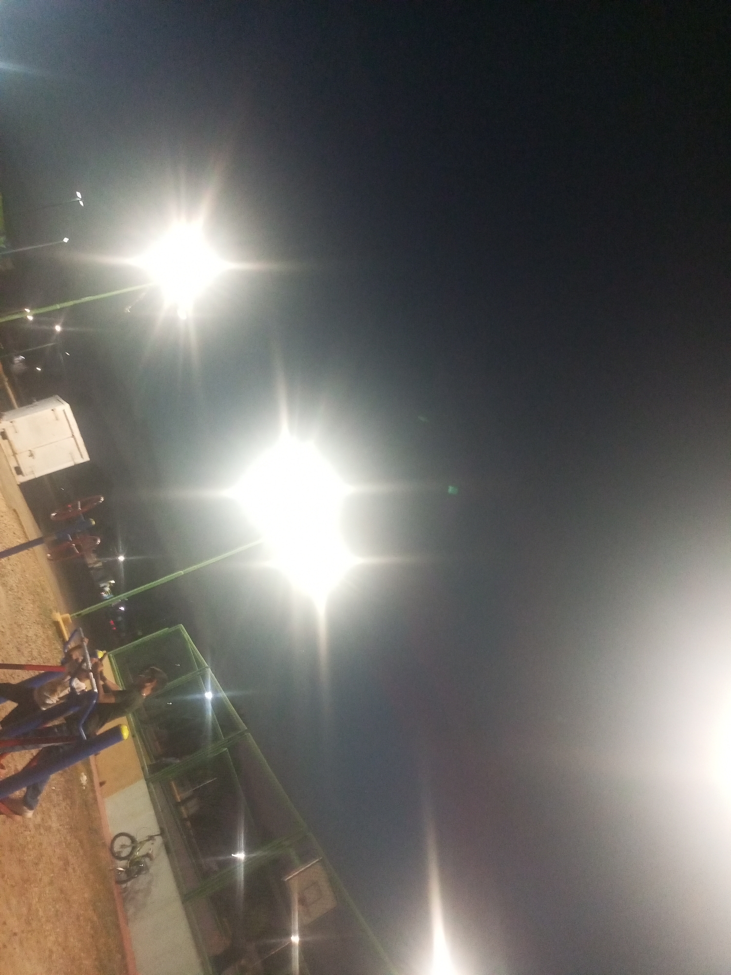 The image shows an outdoor scene at night, possibly a playground or recreational area, illuminated by bright overhead lights. The location appears to be in Juan Griego, Venezuela.
Subjects and Objects:
* Several tall light poles illuminate the area.
* There are several people, but their features are not very clear.
* Playground equipment, likely swings, seesaw, and a jungle gym, can be identified.
* A building or structure is visible.
* A bicycle is laying down in the playground
* The ground appears to be dirt or gravel.
Actions and Interactions:
* People can be seen playing in the playground.
Environment:
* It is night time.
* The area is artificially lit.