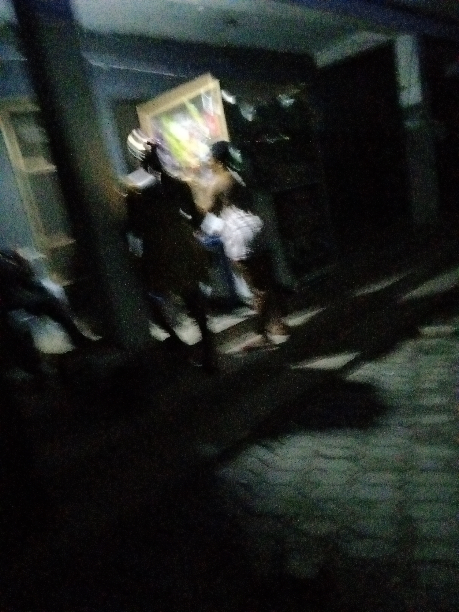 The image is blurry and taken at night. It depicts a group of people walking on a paved surface with shadows cast by an unknown light source. One person appears to be carrying a rectangular object, possibly a framed picture or small piece of furniture. The location is possibly Ado, Nigeria, but this is unconfirmed given image quality.