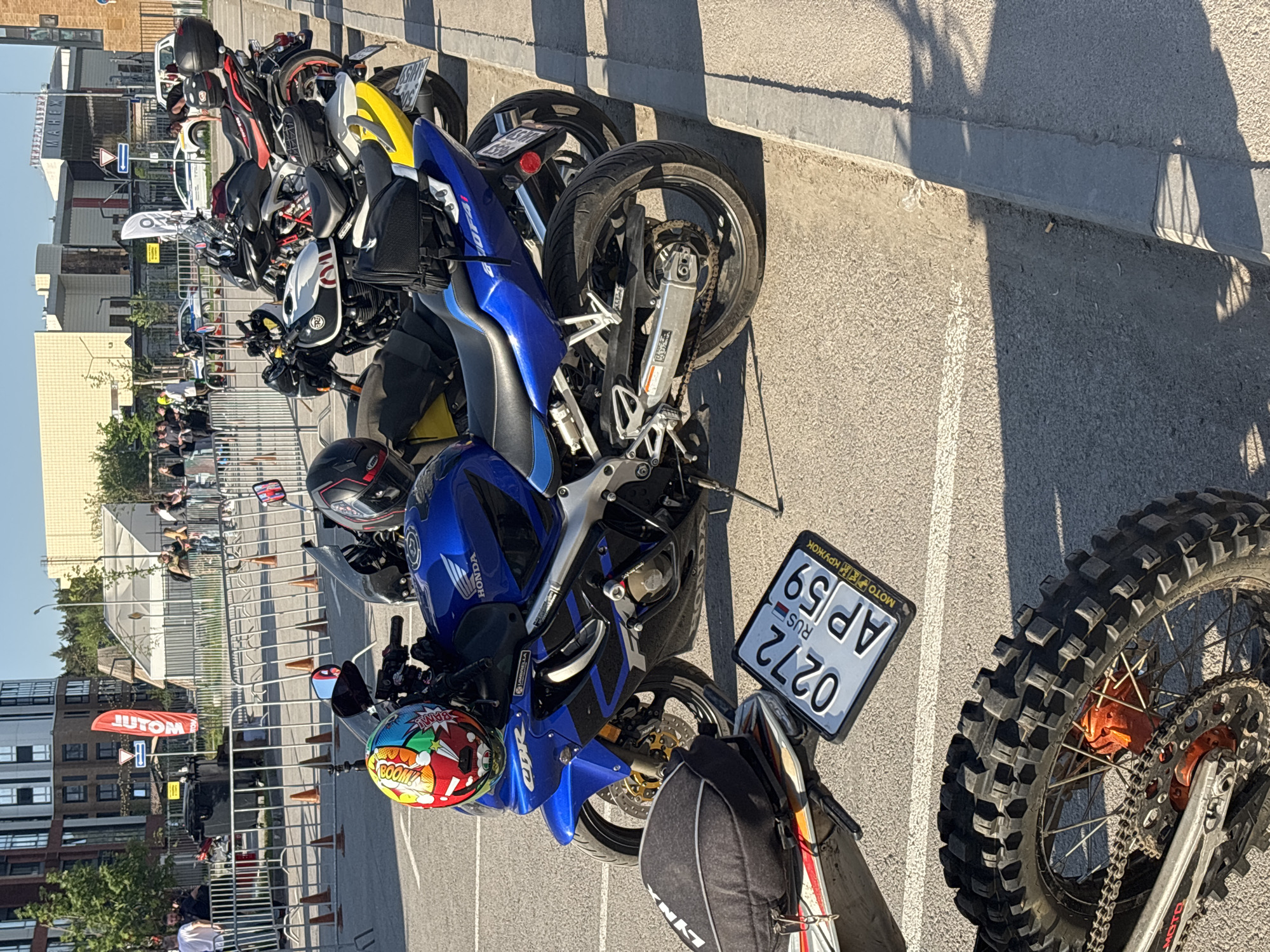 The image shows a row of parked motorcycles in Perm, Russia. The foreground features a blue Honda CBR motorcycle with a colorful helmet on the handlebars, a black bag, and a license plate that reads "0272 AP 59 RUS." Next to it is a motorcycle tire with large treads. Behind the Honda CBR are several other motorcycles parked in a row, and in the background, there's a group of people standing near a metal fence and some orange cones. Buildings are visible in the background as well, and a red banner with "JUTOM" written on it.