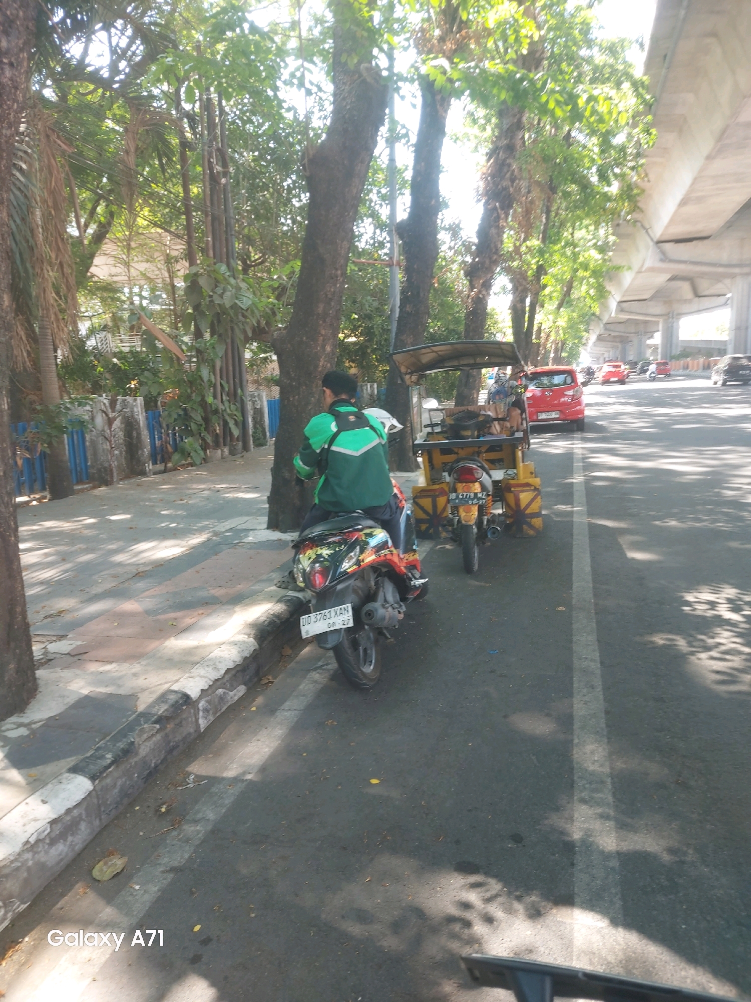 In Makassar, Indonesia, on a sunlit day, a man wearing a green jacket is seen sitting on a motorcycle. The motorcycle is parked on the side of a road, near a sidewalk with a decorative blue fence and trees. The motorcycle has a license plate that reads "DD 3761 XAN". Beside the motorcycle is a yellow three-wheeled vehicle, possibly a pedicab or a modified motorcycle with a passenger carriage. Further down the road, several cars are visible, including a red sedan and other vehicles moving under a highway overpass. The road is asphalt and has a white line marking a lane. The overall scene depicts a typical urban streetscape in Makassar, with a blend of personal transportation and public infrastructure.
