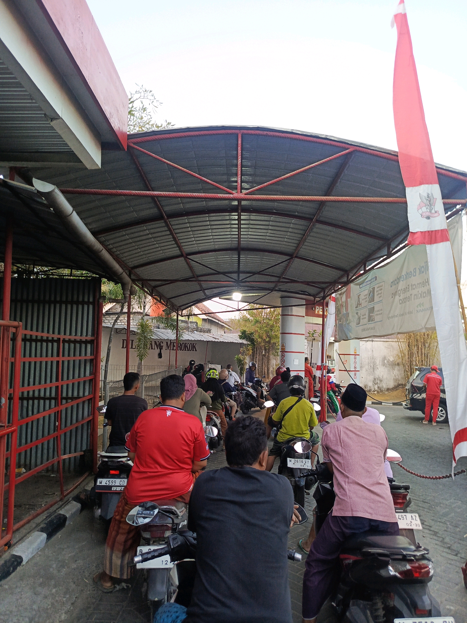 The image depicts a queue of motorcycles at a gas station, possibly located in Gladak Anyar, Indonesia. The scene is covered by a metal roof supported by red and white poles, typical of many gas stations in the region. A prominent Indonesian flag, red and white, is displayed on the right side of the image.
In the foreground, several people are seated on motorcycles, facing away from the camera, waiting in line. The individuals are dressed in casual clothing, with some wearing traditional Indonesian attire like sarongs. The motorcycles are lined up along a paved surface, with a red and black striped curb visible on the left.
Further back, more motorcycles and people are visible, as well as gas pumps and a car being serviced. A banner with text in Indonesian, likely advertising fuel or promotions, hangs across the covered area. The background shows a building with a "DILARANG MEROKOK" (No Smoking) sign and some vegetation. The overall atmosphere suggests a routine day at a busy gas station.