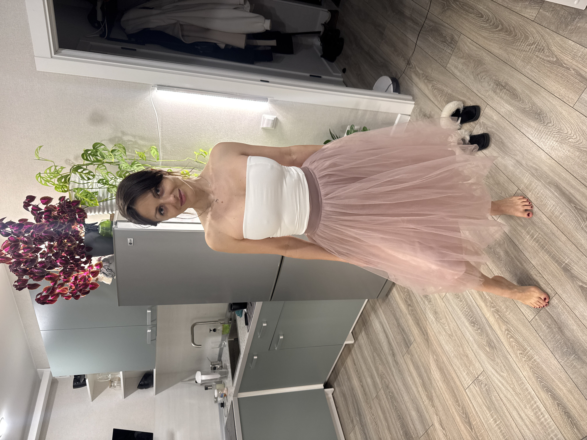 A medium shot captures a woman in a white strapless top and a blush-colored tulle skirt, standing in what appears to be a kitchen. The kitchen features gray cabinetry, a stainless steel sink, and light-colored wood-patterned flooring. Behind the woman, a refrigerator and various plants are visible, including one with purple leaves. The woman is barefoot, and a pair of black and white slippers are on the floor nearby. The location of the scene is Murino, Russia.