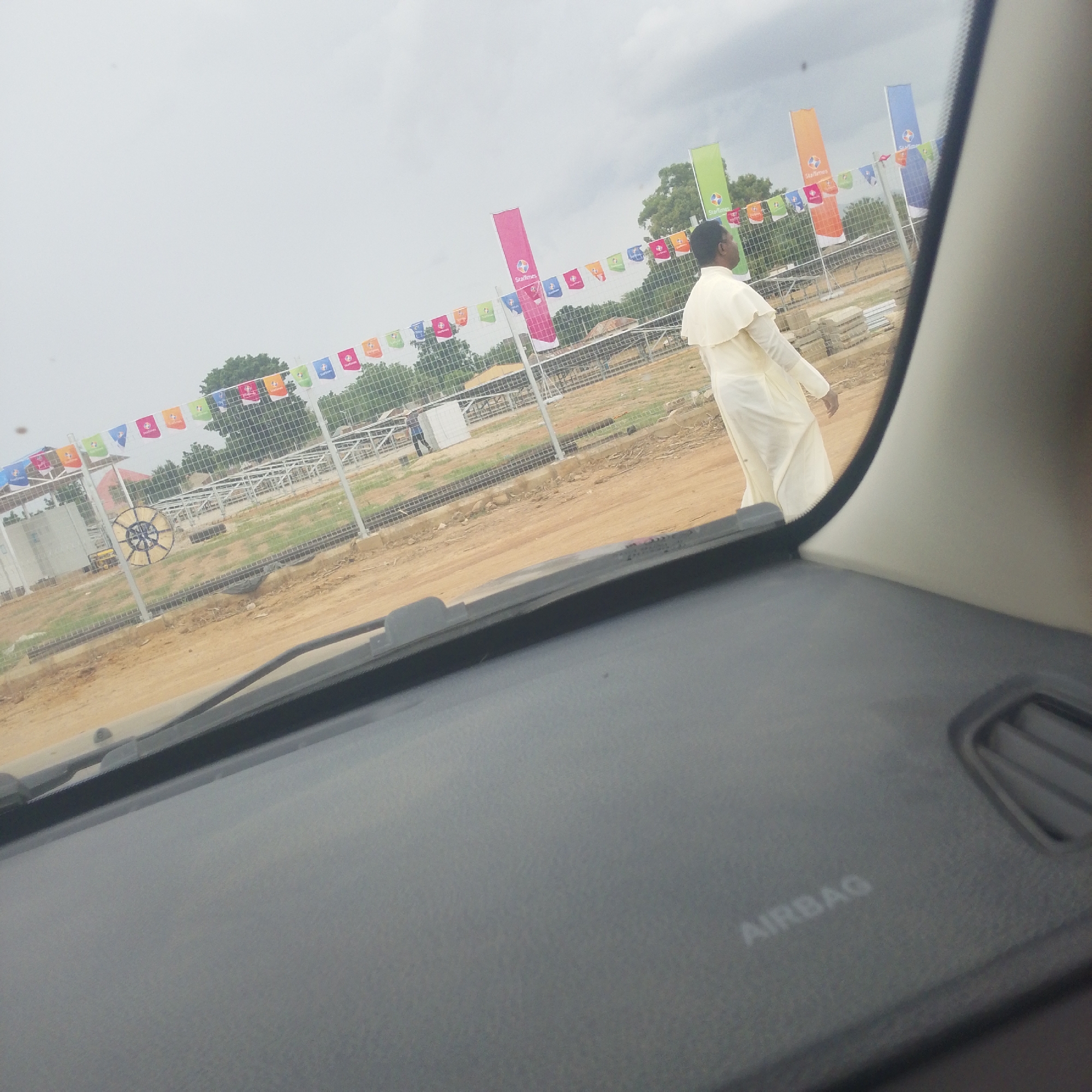 The image was taken in Wuro Ngangandi, Nigeria, showing a construction site with multiple colorful flags and banners hanging from a fence. A person dressed in a cream-colored robe is walking away from the camera on a dirt path. In the foreground, the dashboard of a vehicle is visible, with the word "AIRBAG" imprinted on it. The sky is overcast, and trees can be seen in the background. The construction site appears to be in an early stage, with piles of building materials present.