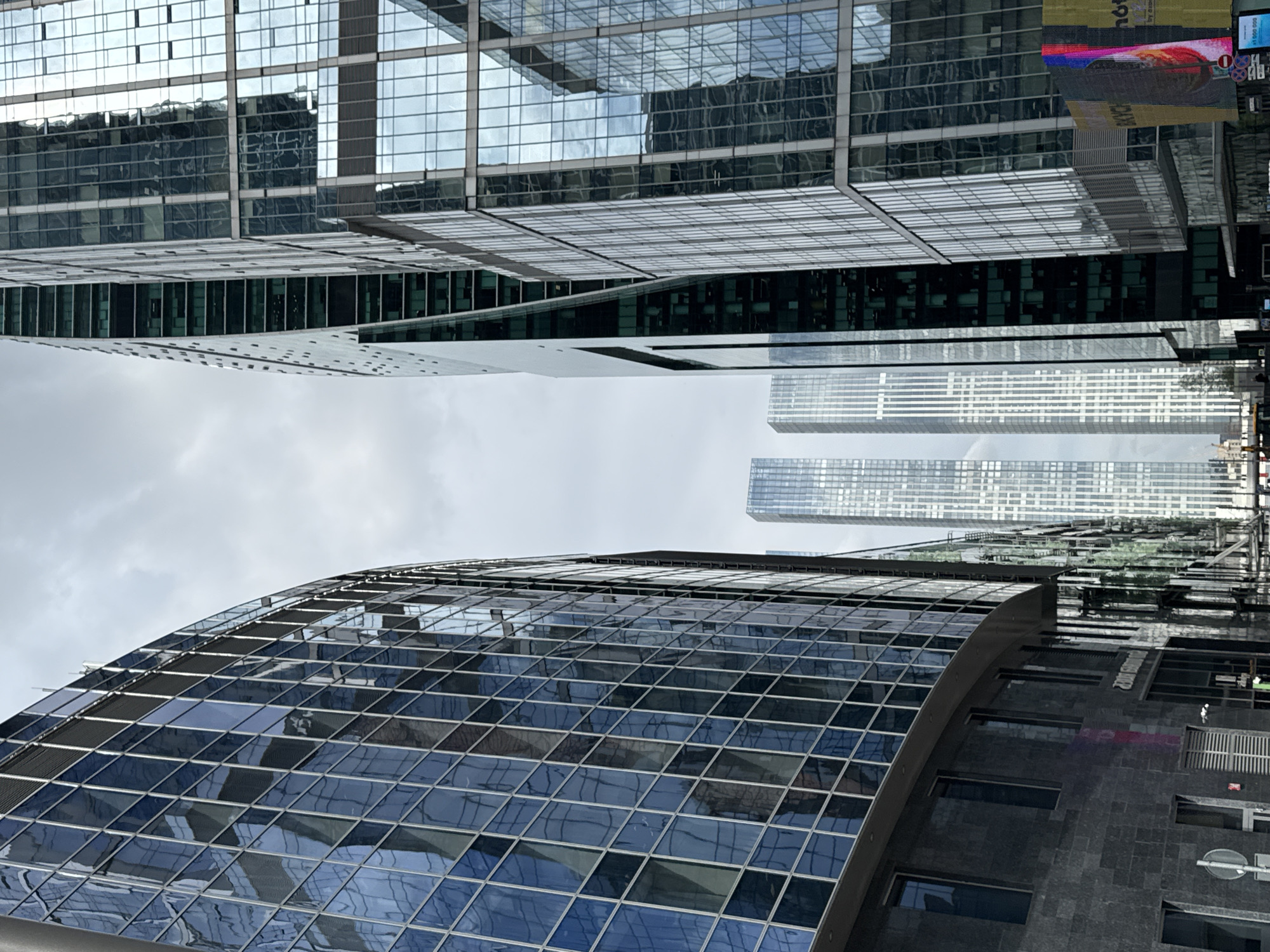 The image shows a low-angle view of buildings in what appears to be a modern urban environment, with a focus on architecture featuring glass and steel. The foreground displays a curved building with a grid-like pattern of glass panels, reflecting the sky and surrounding structures. In the background, there are taller buildings with a rectangular design, also reflecting the sky and other buildings. The sky is overcast, suggesting a cloudy day. The image is rotated in the display, so orientation is not immediately apparent. The location, Khimki, Russia, is given.