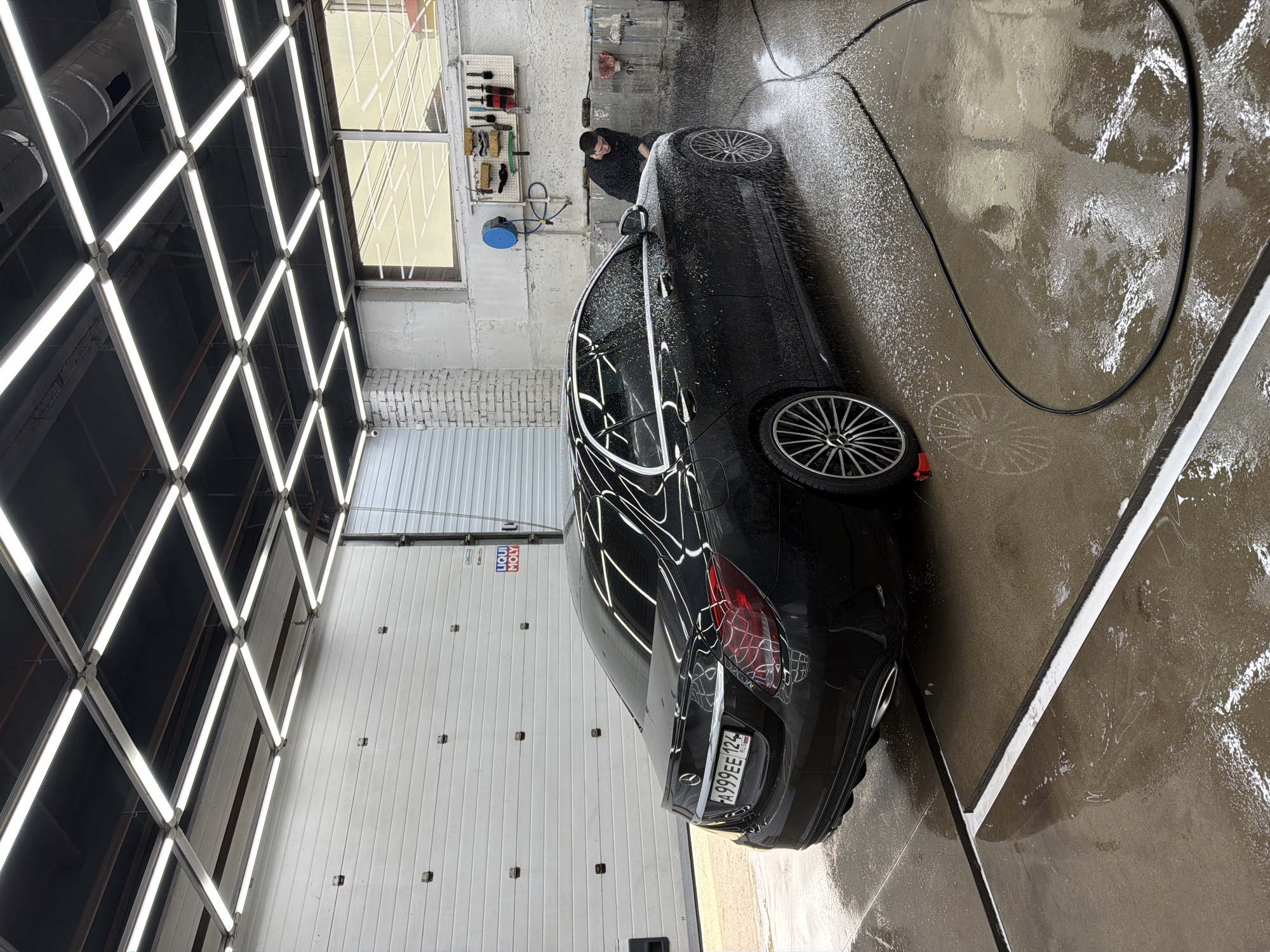 The image shows a black Mercedes-Benz car being washed at a car wash in Krasnoyarsk, Russia. A person is seen cleaning the car near the front left wheel. The car's license plate number is A999EE 124. The interior lighting consists of a grid of fluorescent lights. The car wash bay has a white metal wall with a Liqui Moly logo. The floor is wet with soap and water. A black hose is visible on the floor. Various cleaning tools are mounted on the wall. A window is visible near the ceiling.