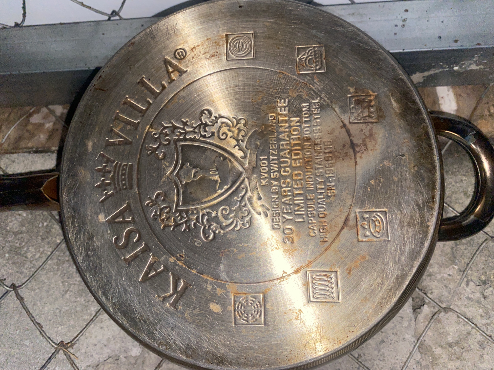 The image shows the underside of a stainless steel frying pan with the brand name "KAISA VILLA" embossed on it. The pan has various symbols and text indicating it is "KV001 DESIGN BY SWITZERLAND 30 YEARS GUARANTEE LIMITED EDITION CAPSULE INDUCTION BOTTOM HIGH QUALITY STAINLESS STEEL SN-1968116". There are also several small square icons with embossed symbols around the rim of the pan. The pan appears to be somewhat worn and has some rust or discoloration. It is photographed from above, and part of a chain-link fence is visible in the upper left corner of the image. The background is out of focus and appears to be a rough, textured surface, possibly concrete or stone. The overall lighting suggests it is an indoor or shaded outdoor environment. There is no indication of any specific location within Davao City, Philippines.
