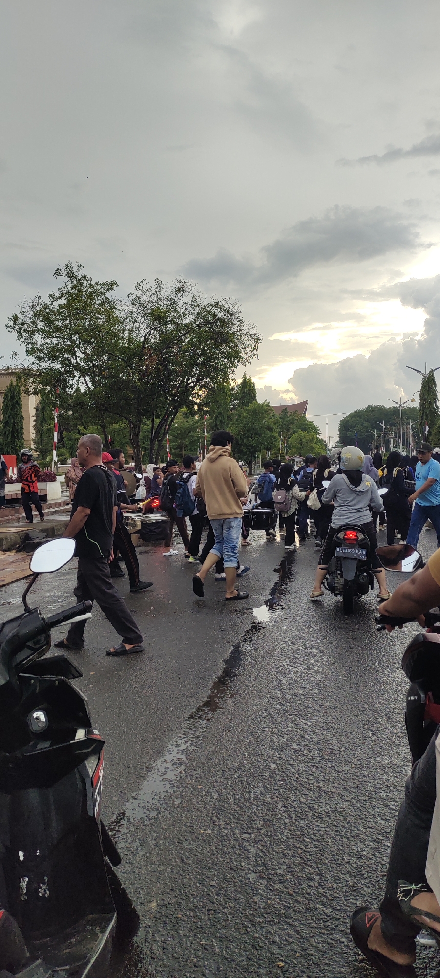 The image depicts a busy street scene in Lhokseumawe, Indonesia, likely near the Baiturrahman Grand Mosque due to the presence of a large crowd and a building that resembles a mosque in the background. The sky is overcast with dramatic, dark clouds, suggesting either the aftermath of a storm or an approaching one. The wet asphalt road indicates recent rainfall, with puddles reflecting the dim light.
A diverse group of people, predominantly young, are gathered in the street. Many are walking, some appearing to be moving with a sense of purpose, while others seem to be observing. There are several motorcycles present, some parked and one in motion, with a rider wearing a helmet. A person in the foreground is seated on a motorcycle, with their arm extended, possibly taking the photograph.
Among the pedestrians, a man in a black t-shirt and dark pants is walking towards the left of the frame. Another individual in a beige hoodie and light blue jeans is crossing the street. A group of people with backpacks and some carrying what appear to be musical instruments, possibly drums, are visible in the middle ground, suggesting a procession or a performance. The general atmosphere appears to be one of communal gathering or activity, with a sense of movement and interaction among the individuals and vehicles. The surrounding environment includes trees and buildings, typical of an urban setting.