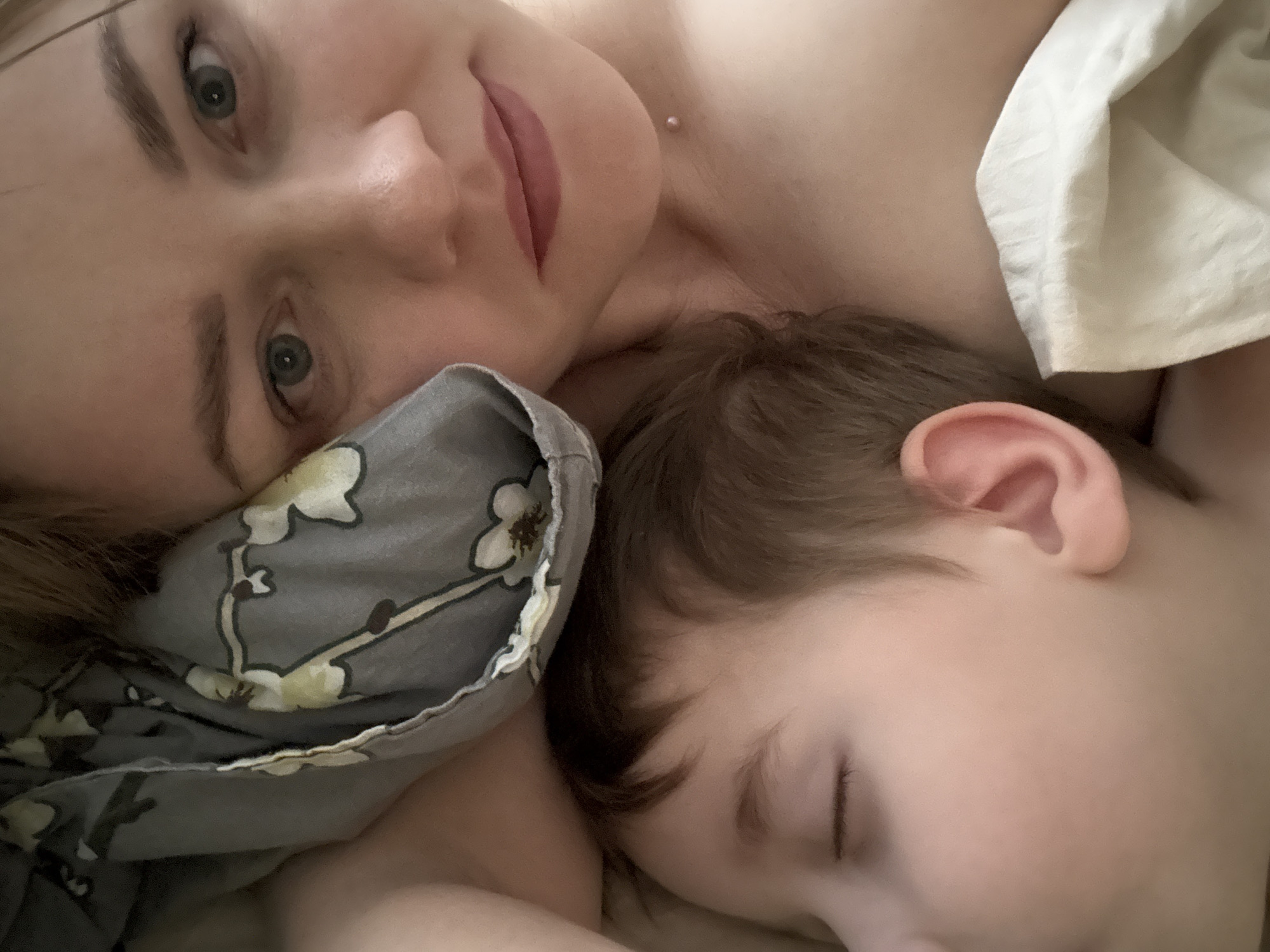 A close-up photograph shows a woman and a young child lying down, possibly in bed. The woman is awake and looking at the camera. The child appears to be asleep with their eyes closed. They are both lying on bedding, including a pillow with a floral pattern. The location in the city of Chaikovsky, Russia is not discernible from the image.