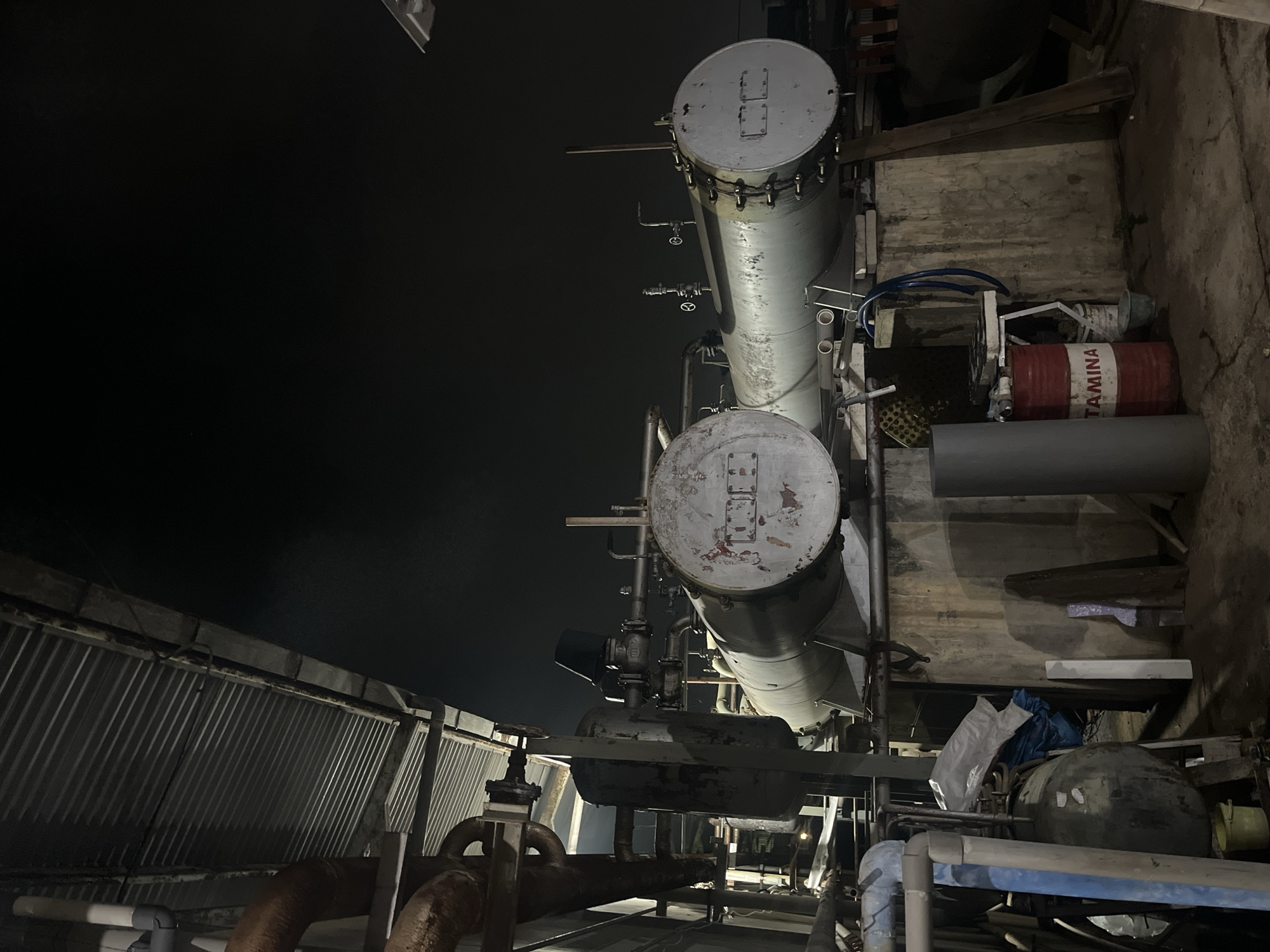 The image depicts an industrial scene at night, likely in Pekalongan, Indonesia, given the presence of a "PERTAMINA" labeled barrel. The foreground is dominated by industrial equipment, including large cylindrical tanks, pipes, and a corrugated metal roof overhead. Two prominent horizontal tanks are visible, one in the center and another to its right, both showing signs of wear and tear. The tank on the left has a faded red and white emblem or text on its end, while the tank on the right has a simpler rectangular marking. Several smaller pipes and valves connect to these tanks.
In the lower portion of the image, a red barrel with white lettering that reads "PERTAMINA" is a key identifier. Adjacent to it, a white bucket and other debris are scattered on the concrete ground. The environment appears to be an outdoor storage or operational area, with concrete flooring and walls visible. The lighting is dim, suggesting it's nighttime, with artificial light sources casting shadows. No people are present in the image. The overall impression is one of an operational industrial site, possibly a fuel depot or processing facility, given the PERTAMINA branding.