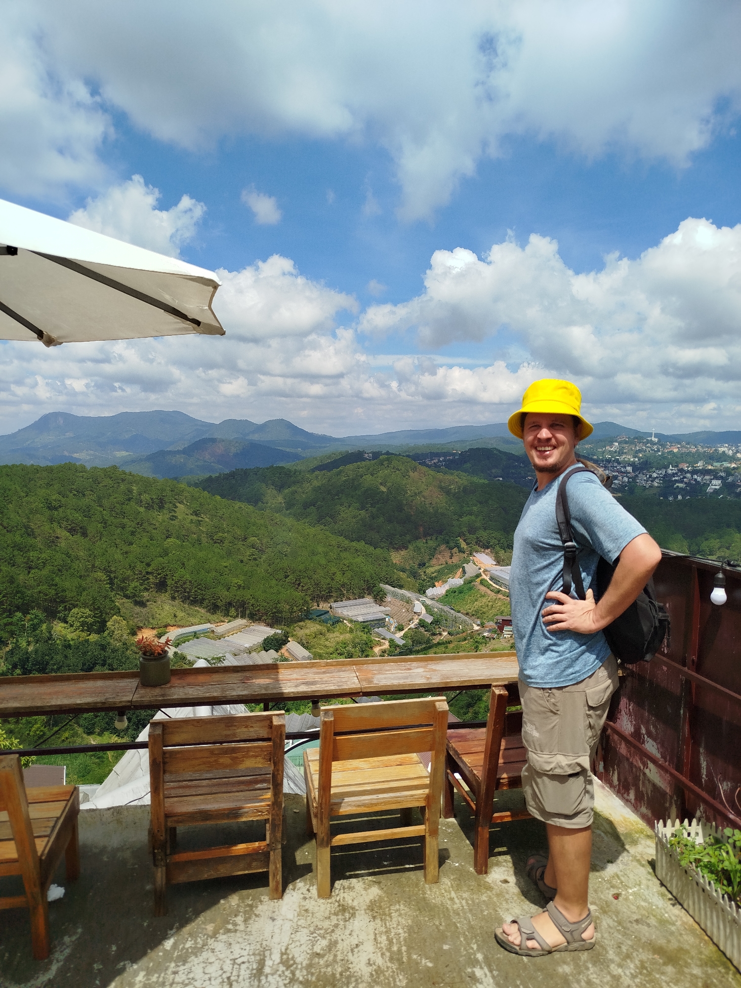 A man with a yellow bucket hat and a blue t-shirt stands on a balcony overlooking a verdant valley with rolling hills and a distant city. He has his right hand on his hip and is smiling at the camera. He is wearing beige cargo shorts and sandals. A black backpack is slung over his left shoulder. In the foreground, there are wooden chairs and tables, suggesting an outdoor seating area. The backdrop features a panoramic view of a lush green landscape dotted with greenhouses and a city sprawling across the hills. The sky is blue with scattered white clouds. The balcony has a concrete floor and a rustic wooden railing. The scene appears to be captured in Xuân Thành, Vietnam, given the context.