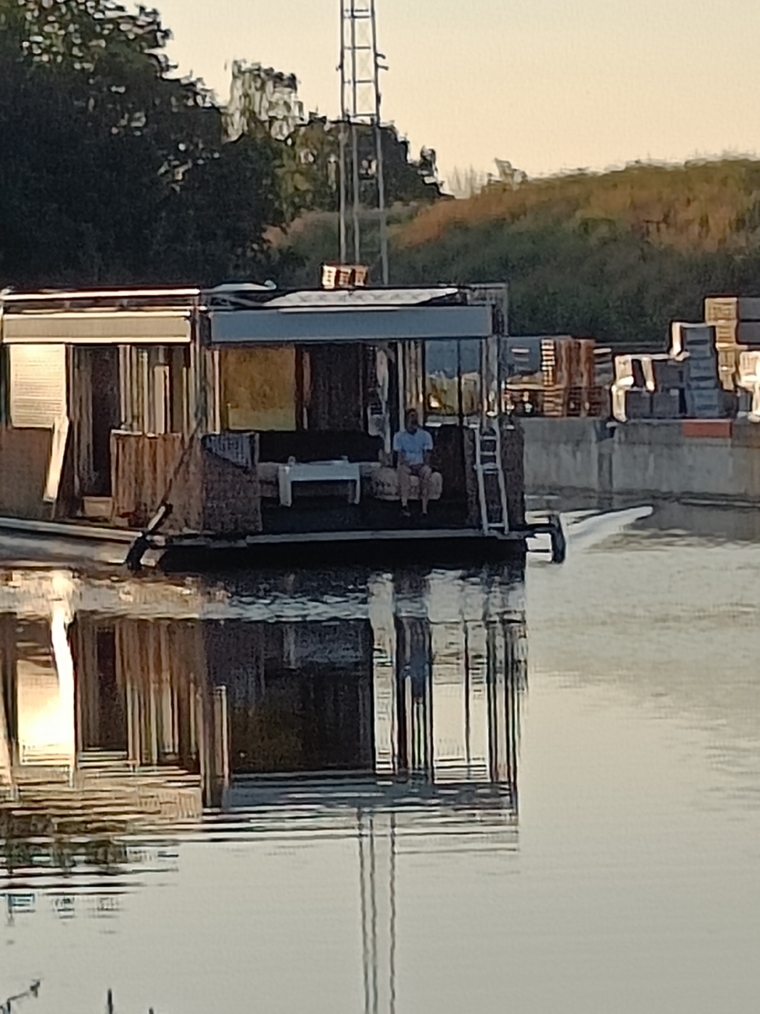 A man is sitting on a couch on the deck of a houseboat. The houseboat has a modern design with large windows and a dark roof. The houseboat is moored in a calm body of water, likely a canal or river, and its reflection is visible on the water's surface. In the background, there are trees, a grassy embankment, and stacked shipping containers. A metal tower structure is also visible in the distance, possibly a radio mast or part of an industrial facility. The scene appears to be set in an industrial or semi-industrial area, with the water providing a contrast to the built environment. The lighting suggests it is either early morning or late afternoon.