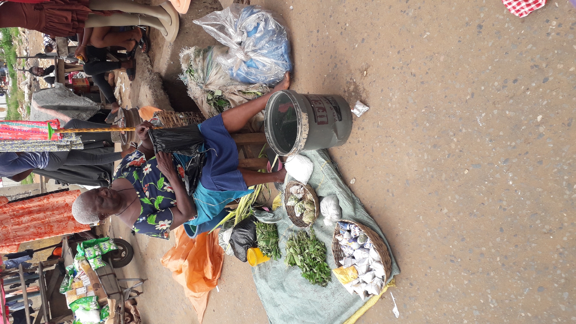 The image depicts a street market scene in Onitsha, Nigeria, with a focus on a woman selling goods. The woman, with gray hair and wearing a floral print blouse and blue shorts, sits on a low stool, her legs stretched out. She is holding a black plastic bag. Beside her, a large gray bucket with some liquid is visible.
The ground is paved with asphalt. Various items are laid out on a green cloth and in baskets. These include bundles of leafy green herbs, some tied with string, and other wrapped items that appear to be packaged goods or produce. There are also woven baskets containing small, wrapped items. In the background, stacks of fabric with colorful patterns are visible, along with other market stalls and people. A cart with bags of what appear to be food items is also present.
The lighting suggests it is daytime, possibly mid-morning or afternoon. The overall impression is a busy, functional market environment. There is a sense of everyday commerce taking place. Some text is partially visible on the gray bucket, appearing as "CCI" in a dark color.