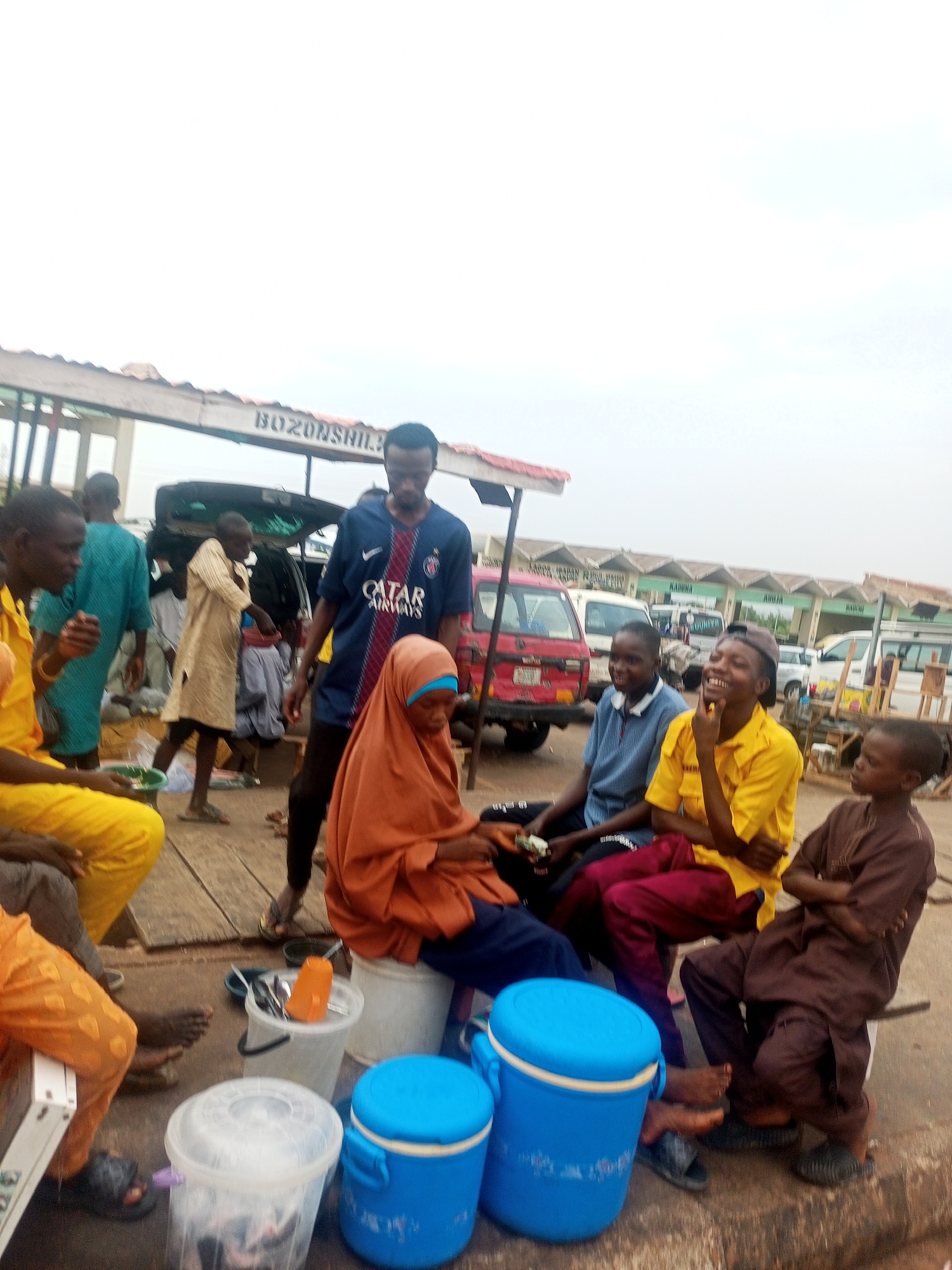 The image depicts a group of people gathered in Gombe, Nigeria. There are men, women, and children present. The scene appears to be an outdoor market or gathering place. The sign "BOZONSHILI" is visible on the structure. One person is wearing a "Qatar Airways" soccer jersey. There are blue ice chests present. The backdrop shows vehicles and structures with signs that read "KADUNA", "ABUJA", and "MAIDUGURI". The sky is overcast.