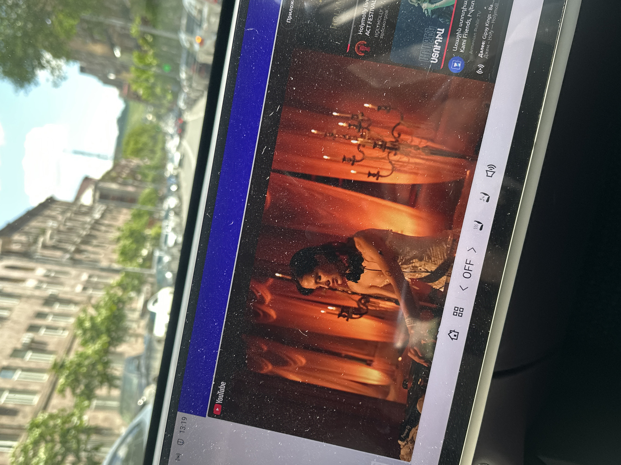 A tablet displaying a YouTube video is angled in the foreground. The video shows a person with dark hair in an ornate, red-lit room, possibly performing or acting. The tablet's screen also displays control icons, time (13:19), app notifications, and some text in Armenian and Russian. Through the car window, a street scene is visible. The environment consists of a building with multiple windows, trees, and other cars. The city is Yerevan, Armenia.