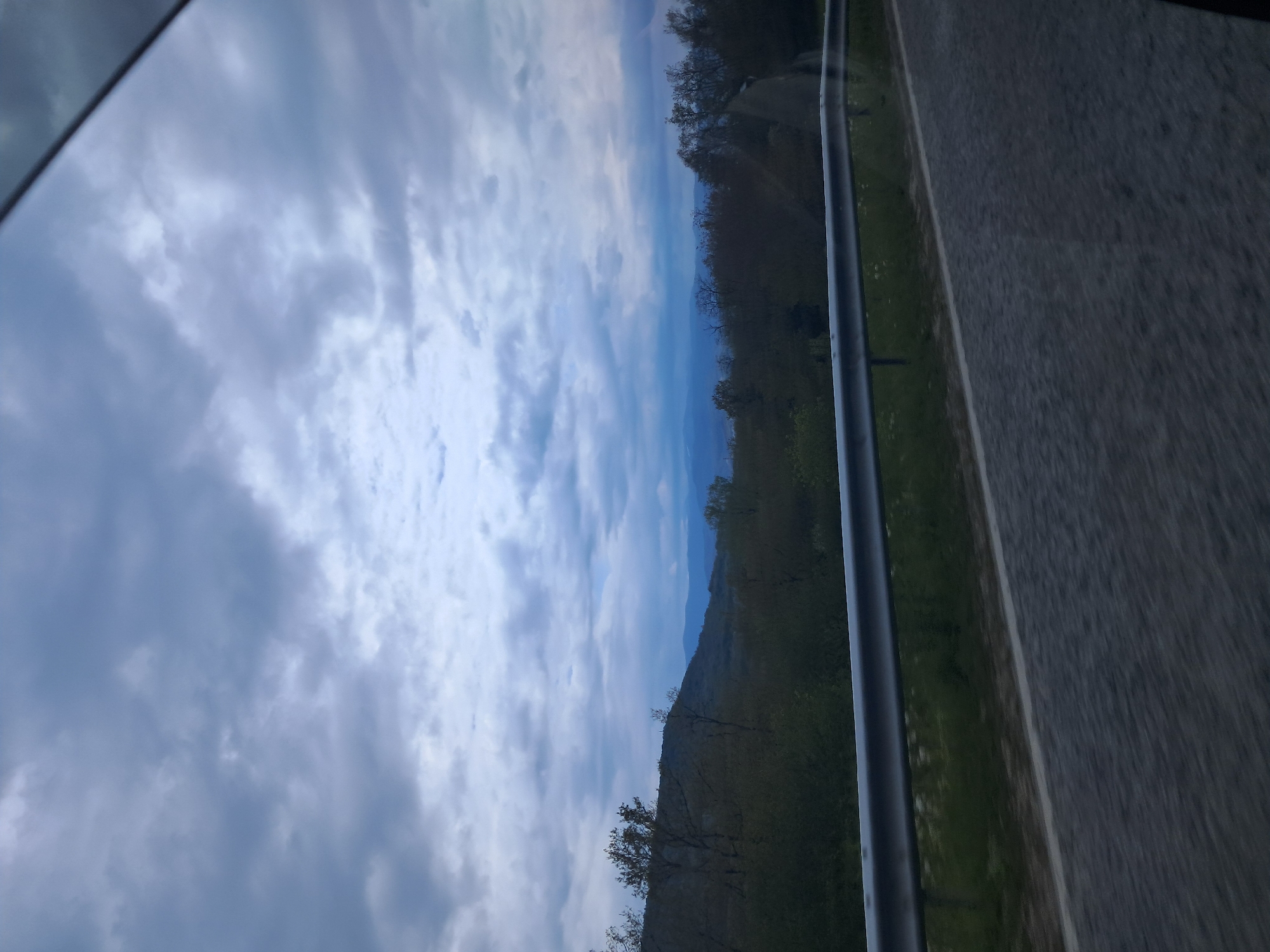 The image depicts a view from inside a vehicle, likely a car, showcasing a landscape in transit. The sky is filled with cloudy formations, occupying a significant portion of the frame. Adjacent to the sky is a view of a mountainous region, partially covered with trees and vegetation. Next to the green vegetation, there is an asphalt road.
The image was most likely taken in a mountainous area, but it's unclear if the picture location is in Хаджийско, Bulgaria.