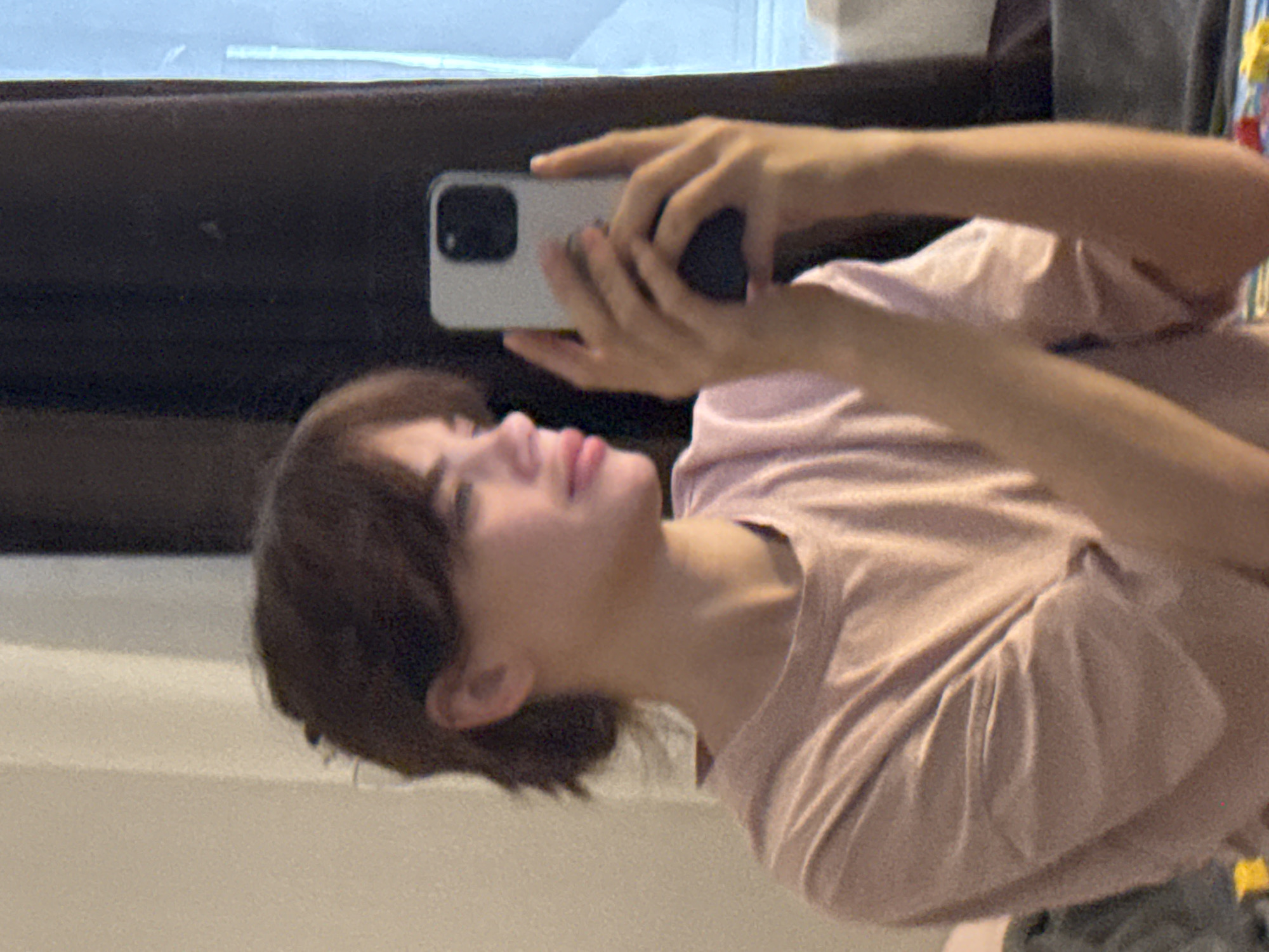 A person with short brown hair, wearing a light pink t-shirt, is holding a white smartphone in their hands and looking up at the screen. The background appears to be a room with a window and a dark surface, possibly a piece of furniture. There is insufficient information to connect the image to Rostov-on-Don, Russia.