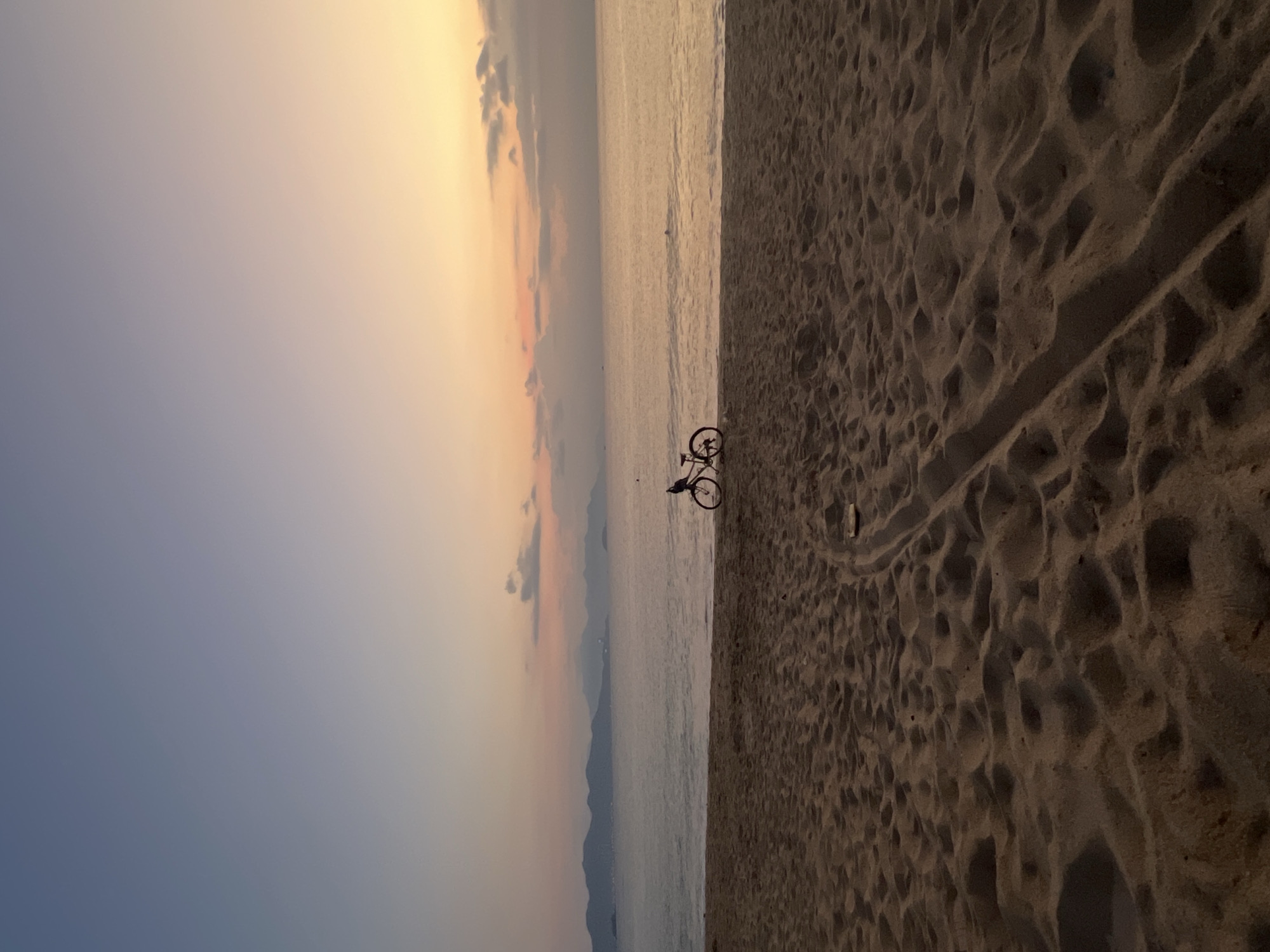 The image depicts a beach scene at dusk in Nha Trang Ward, Vietnam. A person stands next to a bicycle on the shoreline. The beach is sandy, with tire tracks visible. The sea is calm, and the sky is a gradient of yellow, orange, and blue. There are distant mountains visible on the horizon.