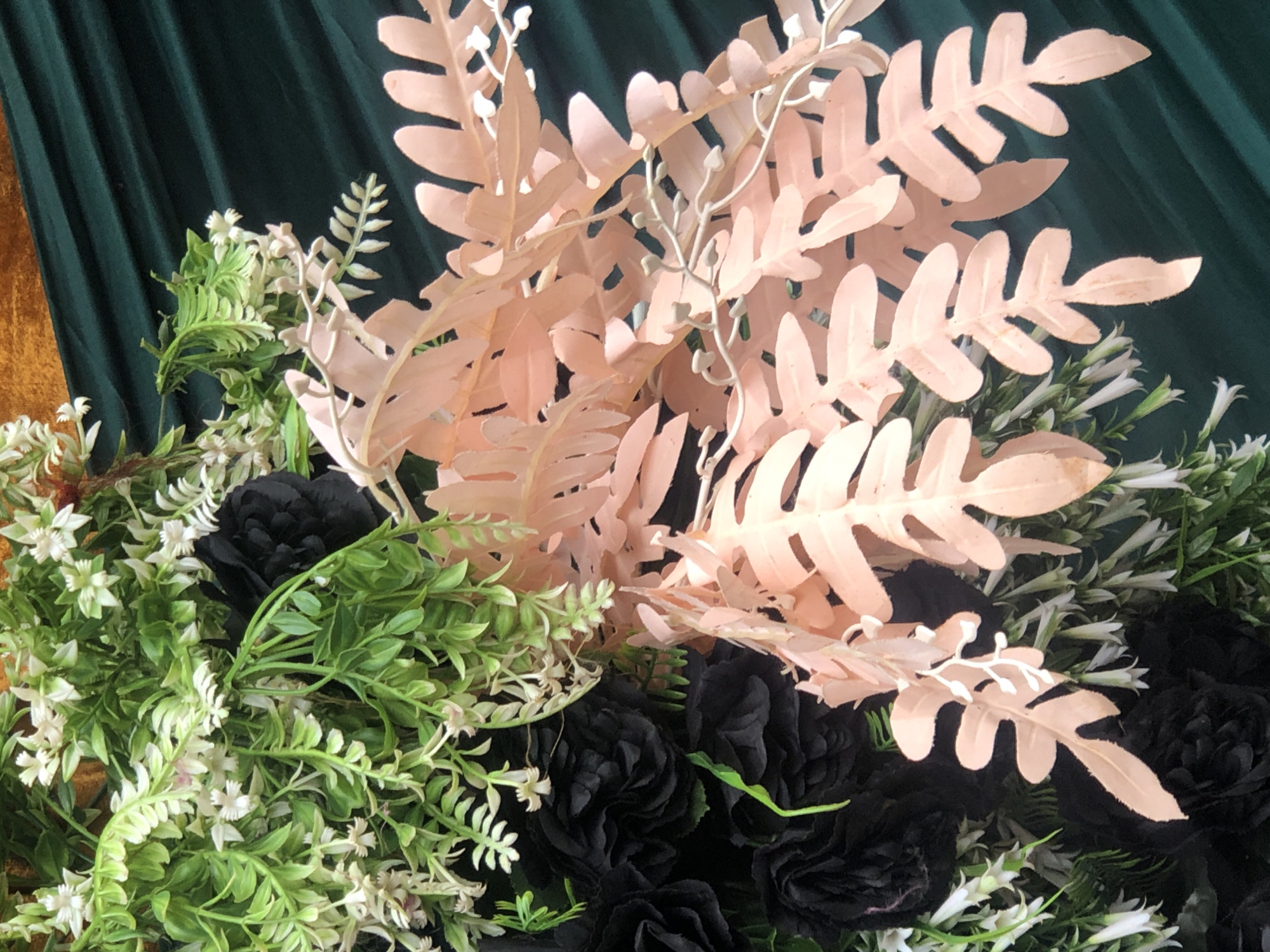 The image features a close-up shot of an arrangement of black flowers, green foliage, and blush-colored fern-like leaves. The background appears to be a dark green, possibly velvet, fabric backdrop. There are no people visible in the image. The location is not discernible from the visual information provided.