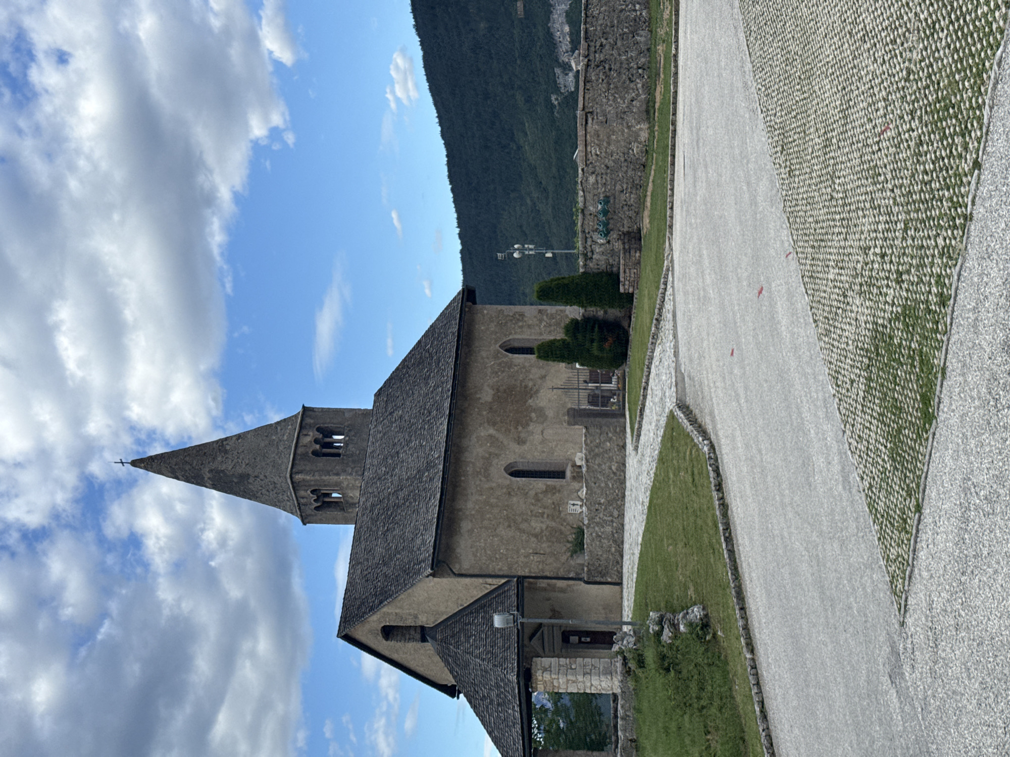 This media file is a photograph taken outdoors in Cavedago, Italy. The scene features a church with a tall, pointed steeple, likely made of stone. The church has a dark, sloped roof and arched windows. Adjacent to the church is a paved pathway, bordered by manicured green grass and stone walls. In the background, a steep, tree-covered mountain dominates the landscape under a bright blue sky with scattered white clouds. The lighting suggests it is daytime, possibly late morning or early afternoon. No people are visible in the photograph. The overall atmosphere is peaceful and serene, highlighting the architecture of the church and the natural beauty of the mountainous surroundings. The image is oriented vertically, with the sky and the top of the church steeple on the left side.