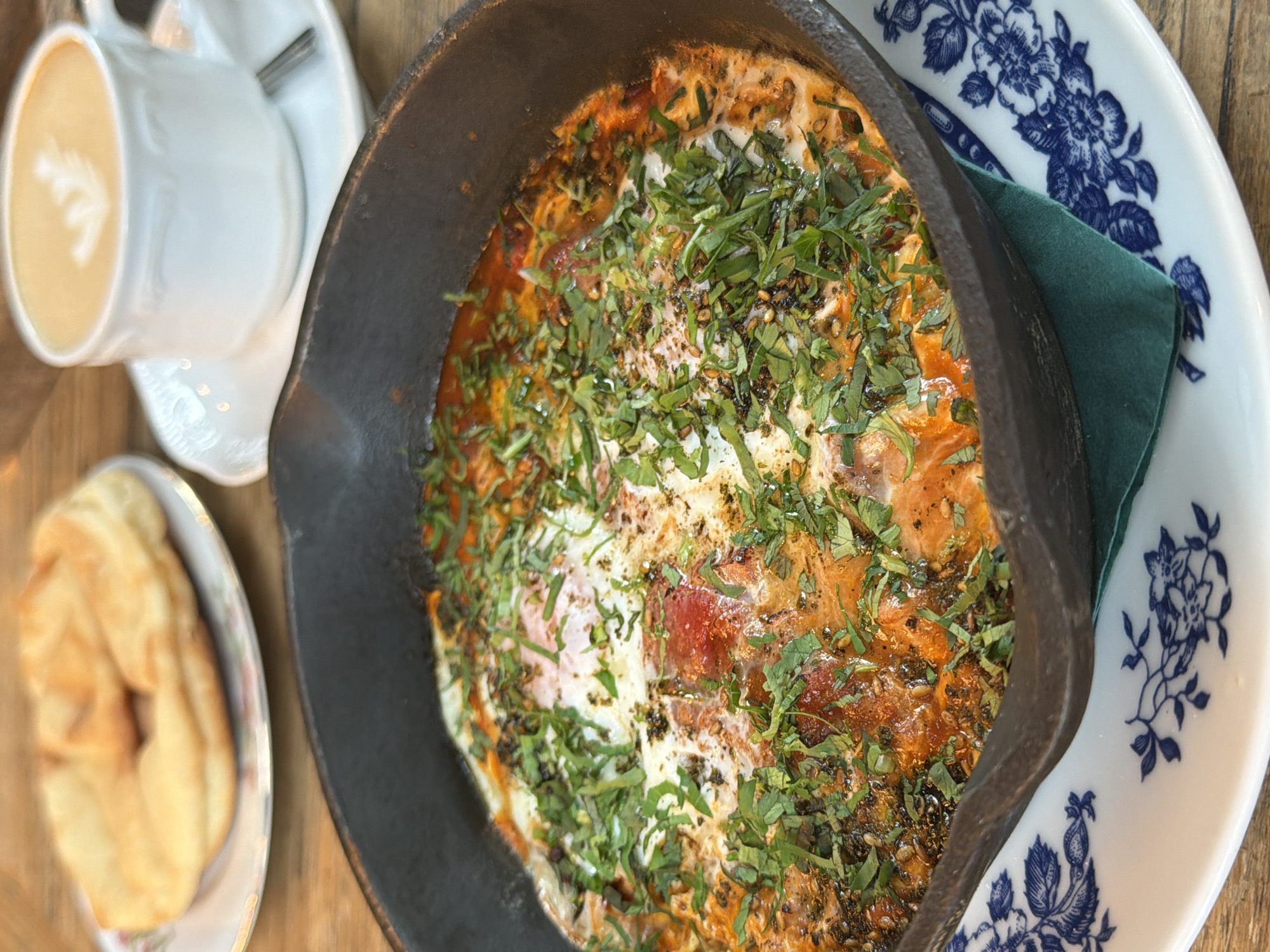 The image shows a close-up of a breakfast dish served in a black cast-iron skillet. The dish appears to be eggs baked in a red sauce, topped with chopped green herbs and spices. The skillet rests on a white plate with a blue floral pattern. In the background, a white teacup with a latte and a piece of bread on a small plate are visible. The setting appears to be a cafe or restaurant, with a wooden table surface. There are no people visible in the image. The overall impression is of a hearty and flavorful breakfast meal.