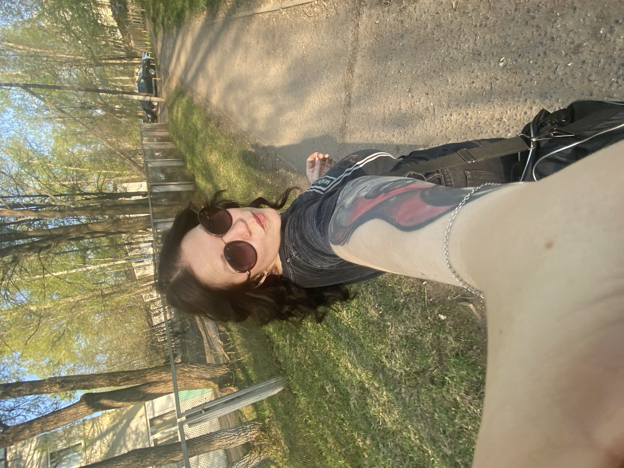 A medium shot of a person with sunglasses facing the camera. The person has dark hair and a visible tattoo on their extended arm. The setting appears to be outdoors on a path or road, with trees and grass visible in the background. A car is parked in the distance, and a building is partially visible on the left side of the frame. Based on the image context, it could be taken in a park or near a residential area. The location is Chaikovsky, Russia.