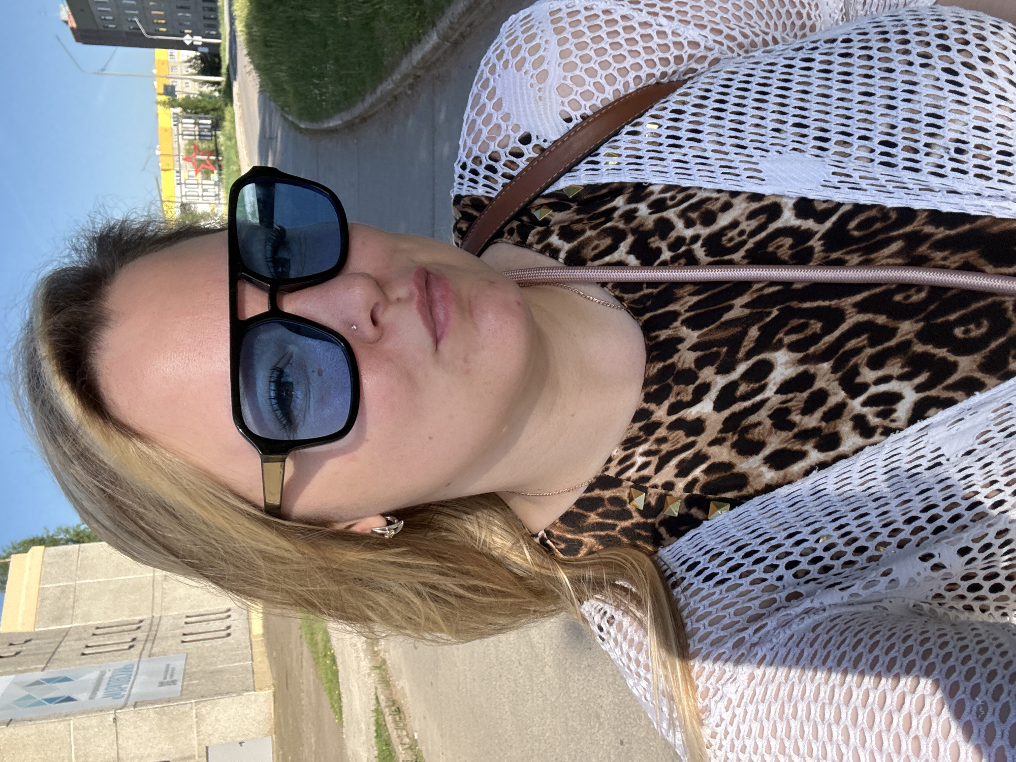 A medium-shot, slightly rotated perspective shows a woman with long, light-blonde hair, wearing sunglasses, a leopard print top, and a white crochet-style cover-up. She also has a nose piercing. The background reveals an urban setting with buildings, a street, and a sidewalk. A building with a prominent red star emblem is visible, aligning with the location of Марковский, Russia.