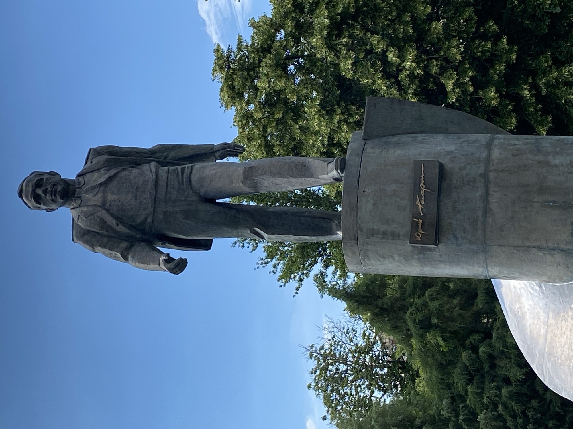 A full-body bronze statue of a man in a suit stands on a cylindrical pedestal. The statue is positioned outdoors under a clear blue sky. The man is depicted with his right leg slightly forward, as if in mid-stride, and his hands are at his sides. He is wearing a suit jacket, shirt, and tie. The pedestal has a plaque on its front with engraved text. Dense green trees are visible behind and around the statue, with a sliver of white cloud in the upper right corner. The statue is partially obscured by a large, curved, reflective metallic sculpture in the lower left corner.