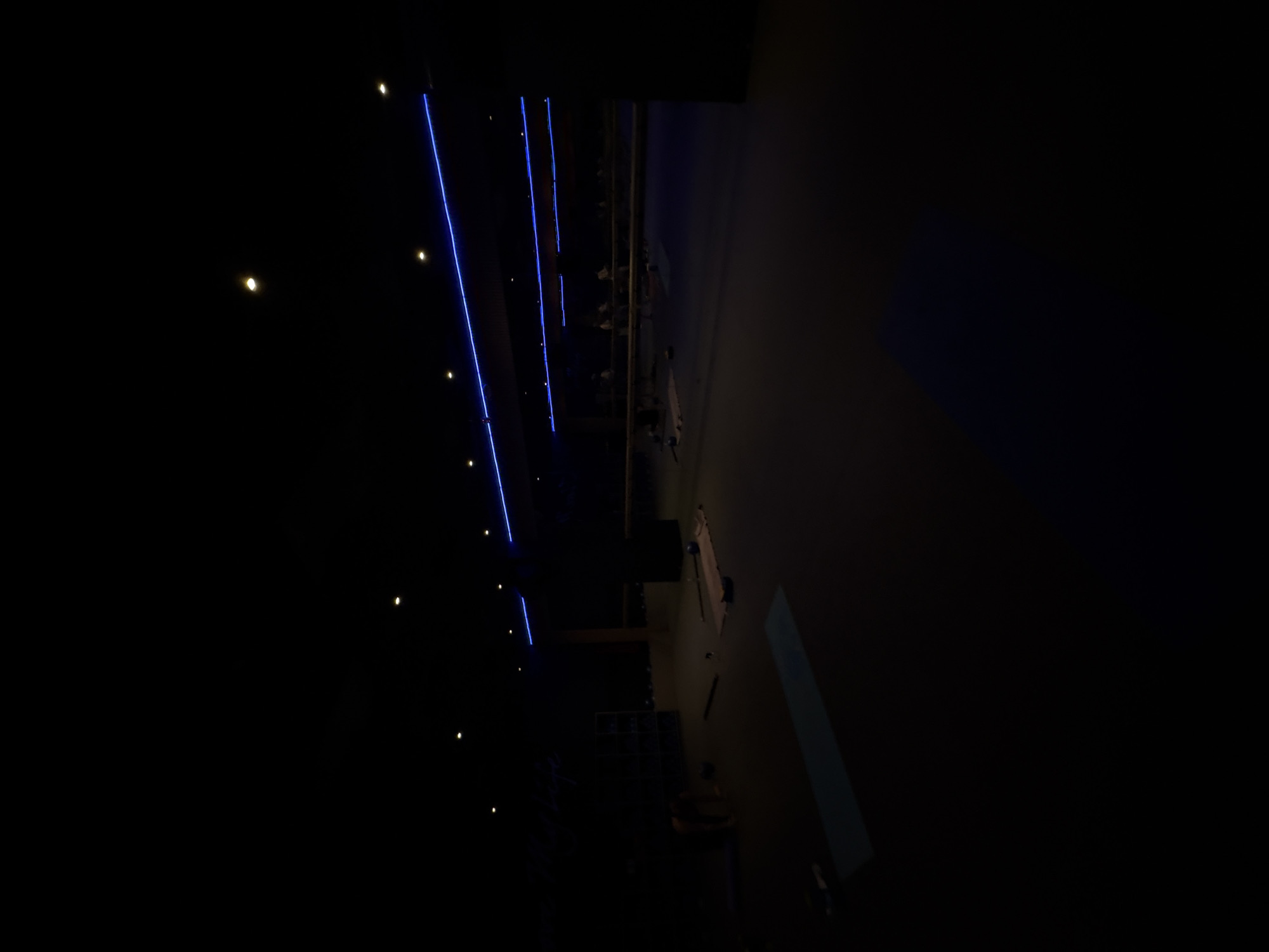 The image depicts a dimly lit interior, possibly a theater or auditorium, located in Mexico City, Mexico. Several rows of seats are visible, with faint figures suggesting people seated. Blue neon lights run along the ceiling, providing a sparse illumination, with occasional small yellow lights interspersed. The environment is generally dark, obscuring many details.