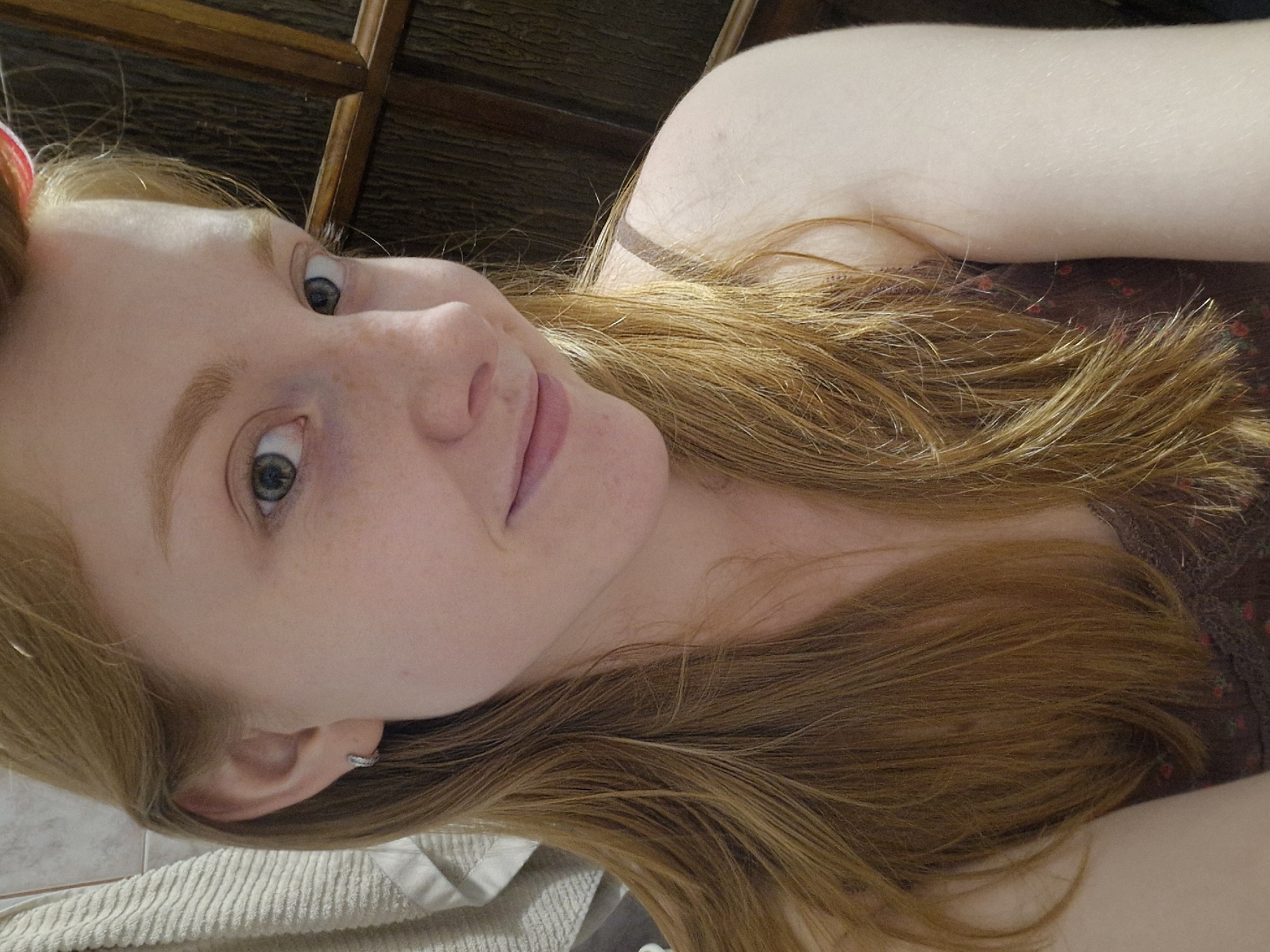 The image is a close-up, selfie-style portrait of a young woman with red hair, fair skin, and freckles. She is wearing a dark-colored top with a floral pattern and a thin strap visible over her shoulder. She has a small silver hoop earring in her left ear. Her hair is long and appears to be casually styled, with some strands falling forward. The background is blurred but suggests an indoor setting with wooden elements, possibly furniture or paneling. A textured towel is visible on the left side of the frame. The overall lighting is soft and appears to be from an artificial source. There is no discernible connection to Yekaterinburg, Russia, in the visual elements of the image.