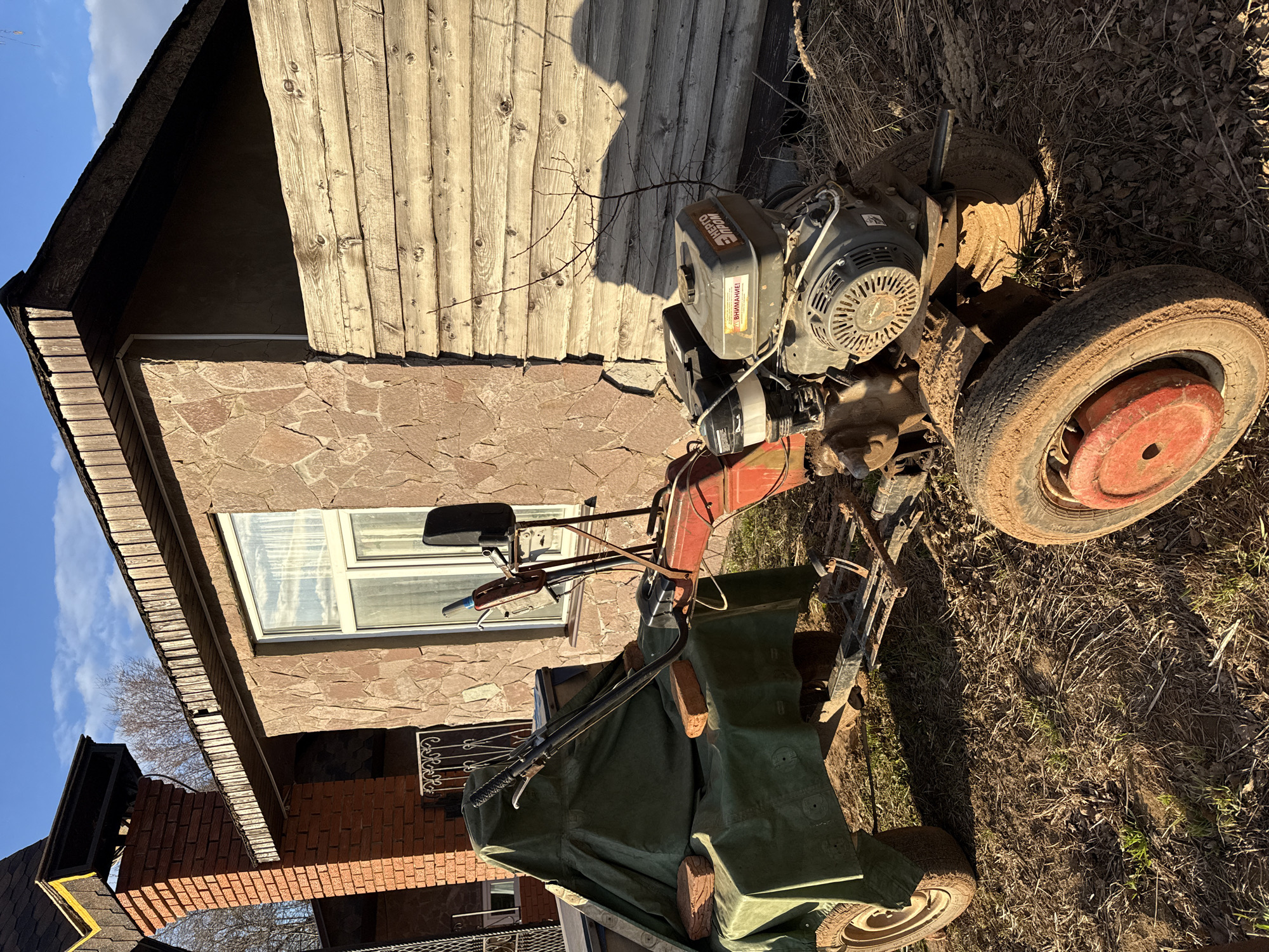 Outside a building in Galevo, Russia, a heavily soiled rototiller sits on dry grass and leaves. The building's facade is partially stone and partially wood. A small window is visible. The rototiller has a red and black engine and muddy tires. Part of the rototiller is covered with a green tarp secured with bricks. The sky is blue and sunny.