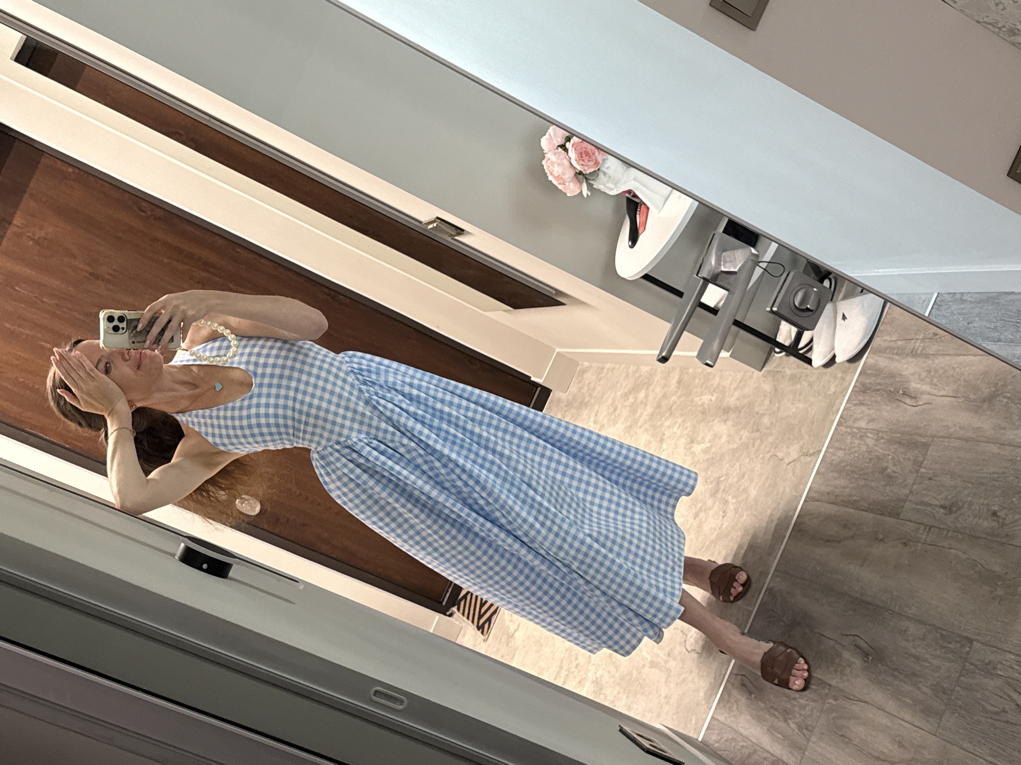 A woman in a light blue and white gingham maxi dress is taking a mirror selfie in what appears to be an indoor setting in Dolgoprudny, Russia. She is smiling and holding a light-colored smartphone up to capture her reflection, with her other hand partially obscuring her face. Her long brown hair is visible. She wears a pearl necklace with a small light blue pendant and a thin bracelet on her left wrist. On her feet, she has brown, open-toed sandals with a woven strap design, and her toenails are painted.
The room features a multi-toned floor, with one section being a light, possibly cream-colored, tile or wood-look surface, and the other a darker, gray-brown wood-patterned flooring, separated by a visible seam. In the reflected background, there's a dark wooden door or paneling, and light-colored walls or trim. A small white round table with a vase containing pink flowers (peonies or roses) is visible, along with a portion of a high chair or stool with a silver frame and white seat. The bright, even lighting suggests it is daytime. No legible text is visible in the image.