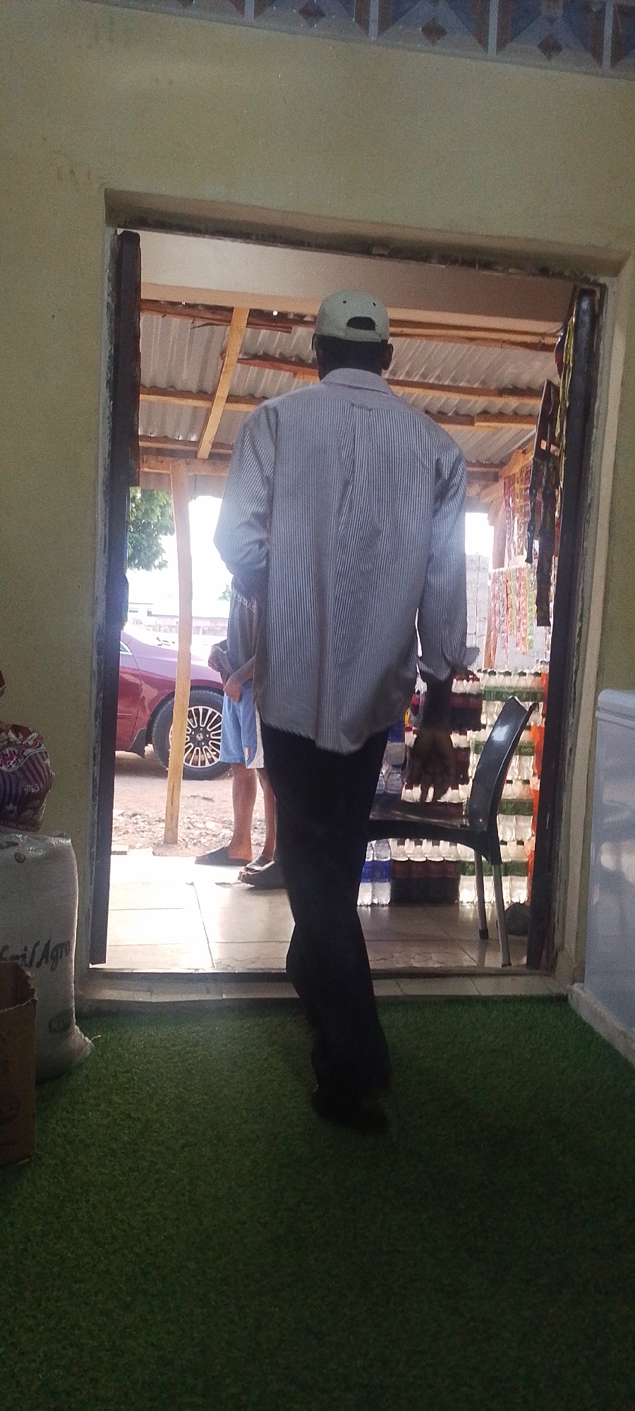 A medium shot depicts an adult male with dark skin wearing a striped collared shirt, dark trousers, and a light-colored baseball cap. He is positioned with his back to the camera as he walks through an open doorway. Beyond him, visible through the doorway, is a small store with shelves stocked with bottled beverages and hanging merchandise. Two individuals are partially visible standing outside the store, one wearing blue shorts and sandals, next to a burgundy-colored car. The floor inside is covered in artificial green turf. To the left of the doorway, a sack labeled "fertilAgro" and cardboard box are present. The setting appears to be Adankolo, Nigeria, based on the cultural context of the scene.