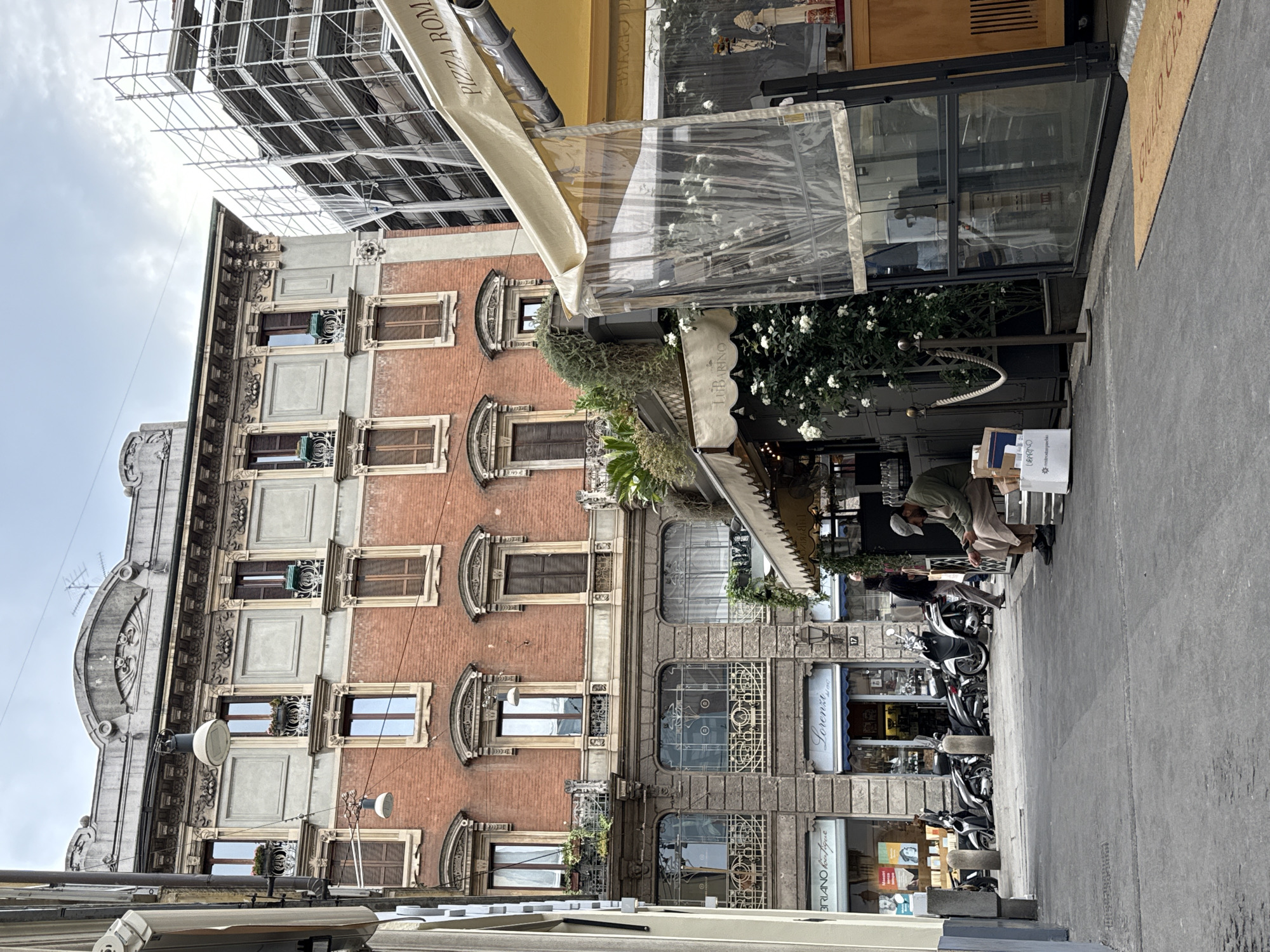 This media file captures a bustling daytime street scene in Milan, Italy, featuring a mix of historic architecture and modern urban life. The perspective looks upward along a paved sidewalk lined with multi-story buildings under a bright, cloudy sky.
On the left, a grand building dominates the frame with ornate architectural details. Its lower floors are constructed of grey stone or concrete, adorned with decorative carvings and large arched windows. Above this, the facade transitions to red brick, featuring several rows of windows, many equipped with closed wooden shutters and small, decorative balconies. Further up, scaffolding covers an adjacent building, indicating construction or renovation.
The right side of the street features several ground-level commercial establishments. A prominent light-colored awning extends over a storefront, displaying the text "PIZZA ROMA". Below it, a transparent plastic enclosure creates an outdoor seating or display area, decorated with white flowers and lush green foliage. Another beige awning partially reads "LIBRINO" or "LIBARNO", and a blue storefront sign below it clearly shows "Lorenza Boutique". A doormat near one entrance faintly displays "GENOVA".
On the sidewalk, a row of scooters is parked. Two people are visible: one individual, dressed in light-colored clothing and a cap, is bending over a box or display near a decorative street barrier with ropes. Another person is partially obscured behind the parked scooters further down the street. The scene depicts a typical vibrant atmosphere of a European city street.