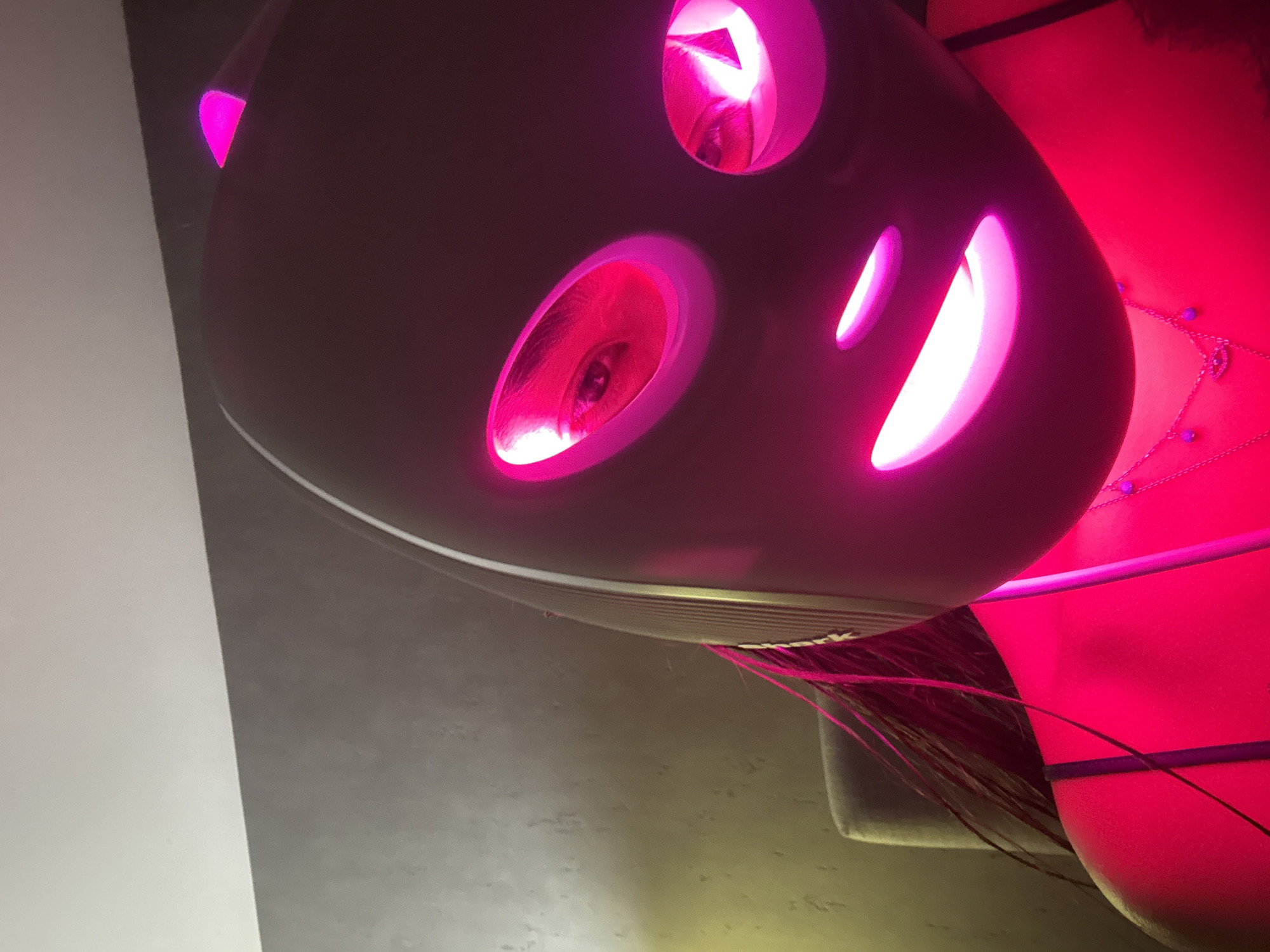An angled, close-up photograph features a dark, elongated object with multiple cutouts, each emitting a strong magenta-pink light. The object's design, with two prominent oval openings and a smaller crescent-shaped one, gives it an almost anthropomorphic, face-like appearance. Within the oval cutouts, reflections of human eyes are clearly visible, indicating interaction or observation by a person.
Faint white text, spelling "Shark," is visible near the bottom edge of the dark object. Extending from its underside are thin, dark, string-like elements, possibly decorative tassels or strands of hair, that reach towards a large, red object positioned to the right. This red object, possibly a bag or piece of furniture, is partially adorned with thin dark lines and a delicate silver-colored chain featuring small purple beads.
The setting appears to be indoors, characterized by low ambient light, which accentuates the vibrant, artificial magenta illumination. A light-colored, possibly white or light grey, vertical surface is present on the far left, contrasting with the predominantly dark and red tones of the rest of the image. The unusual lighting and reflection create an intriguing, almost surreal atmosphere. No specific location cues beyond the object themselves are present in the image.