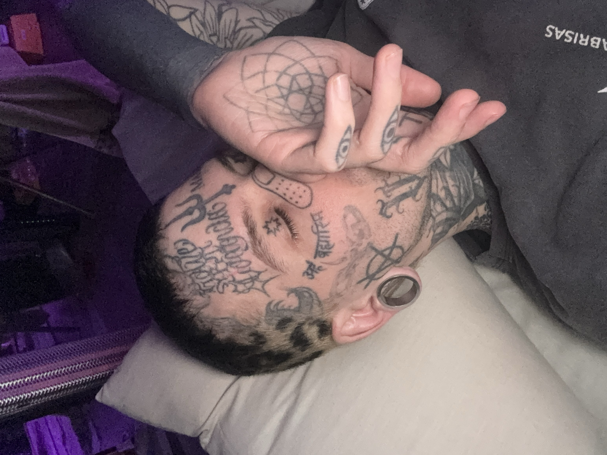 A man with numerous tattoos is lying on a white pillow, seemingly resting. His left hand is placed on his forehead. The man has gauged ears, and a bandage on his forehead. His tattoos feature various designs including geometric patterns and symbols. He is wearing a dark gray sweatshirt. The environment includes bedding and other furniture items in the background. There are no clear indicators of location, let alone Buenos Aires, Argentina.