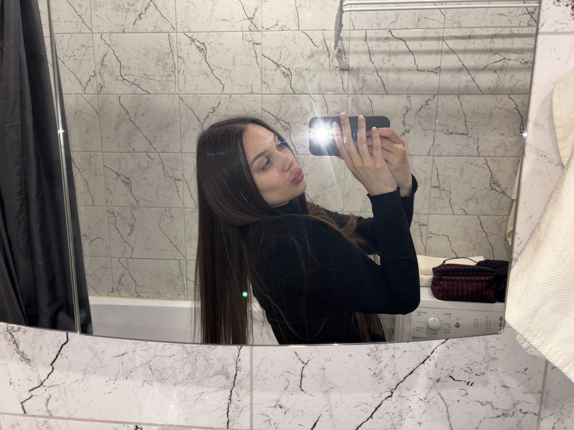 A young woman with long, dark brown hair takes a mirror selfie in what appears to be a bathroom. She is wearing a black long-sleeved top and holds a black smartphone with a bright flash directed towards the mirror, illuminating her face and the surrounding area. Her lips are playfully pursed in a "duck face" expression as she looks towards the phone's camera in the reflection.
The background visible in the mirror consists of white rectangular wall tiles featuring prominent black and grey marble-like veining. To the left, a dark grey or black shower curtain is partially reflected, alongside a glimpse of a white bathtub. On the right side of the reflection, a white washing machine is visible, with a small, rectangular bag adorned with red and black stripes resting on top of it. A white towel rack is mounted on the tiled wall above, and a white towel hangs near the right edge of the mirror. The setting suggests an indoor environment, likely a private bathroom, with the location context being Yekaterinburg, Russia. There is no visible text in the image.
