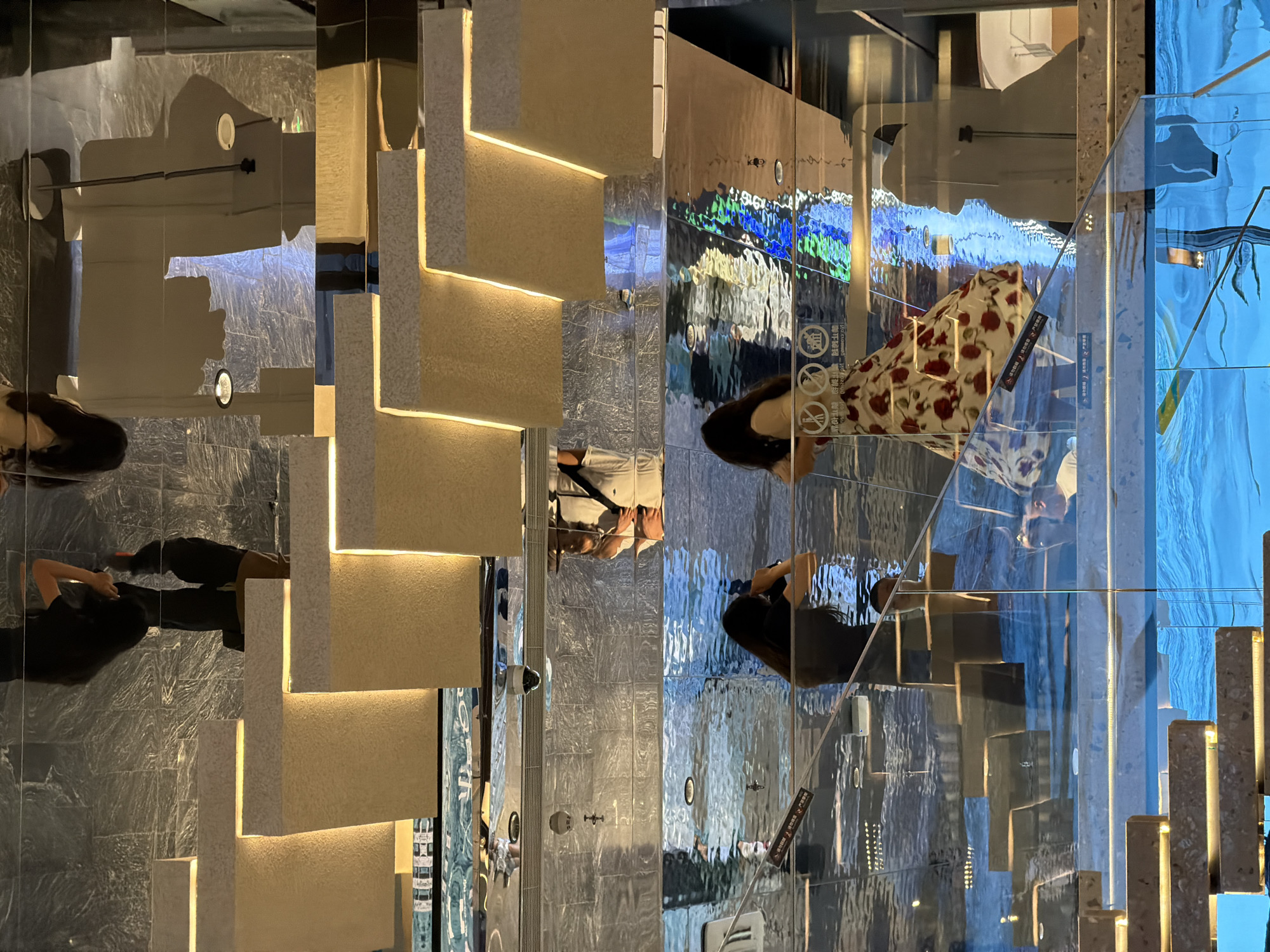 The image shows an interior shot of a modern building, possibly a shopping mall or museum, in Yuzhong District, China. The architecture features illuminated, tiered steps, and a reflective ceiling or floor that creates a distorted mirror effect, showing the scene from an unusual perspective.
In the foreground, viewed from below, are several tiers of the illuminated steps. The steps are made of a light-colored, textured material, with warm LED lights embedded along their edges. Above these steps, the reflective surface dominates, showing upside-down reflections of people and architectural elements.
Two women are visible in the middle ground, appearing in the reflection. One woman, wearing a dark top, is holding a camera to her face, as if taking a photo. The other woman is wearing a floral dress with red roses. They are standing on what appears to be a glass walkway or staircase, which also reflects the surrounding environment.
Further up, in the upper part of the image, more reflections of people and architectural features are visible, including what seem to be hanging lights and ceiling details. The overall impression is one of a highly modern and visually complex space, emphasizing light, reflection, and geometric design. The scene is captured from a low angle, looking upwards into the mirrored architecture.