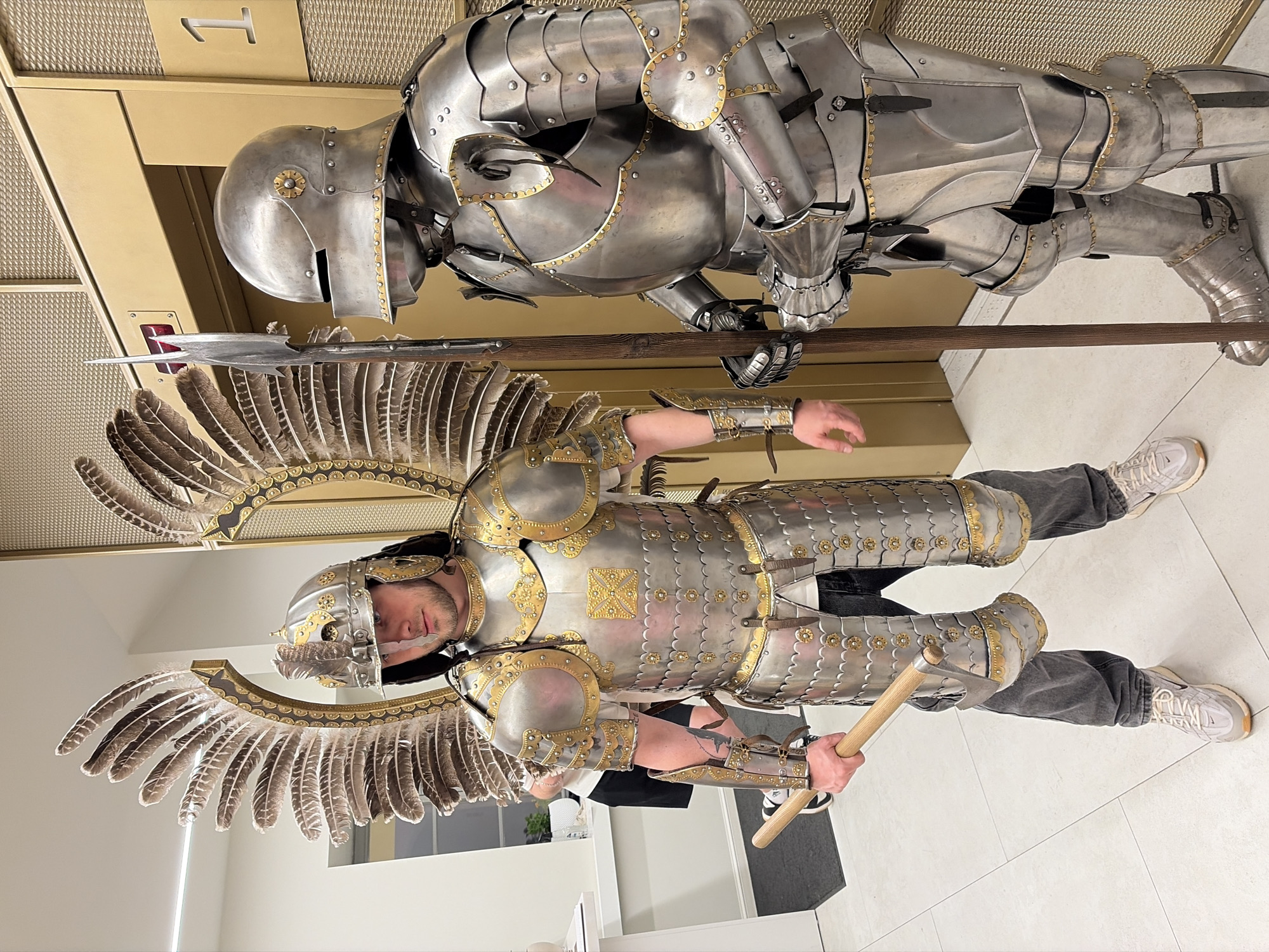 Two men are dressed in full suits of armor and are holding weapons, an ax and a spear. The man on top is not wearing wings, and the other is. The location is indoors in a room with white tiled floors. The exact location in Minsk, Belarus, is not specified.