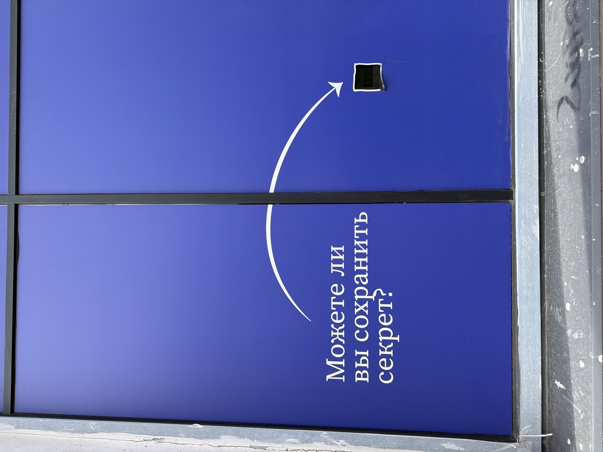 This image captures a vibrant blue sign or panel, likely outdoors in Saint Petersburg, Russia, under bright daylight. The main subject is the blue surface, which features a small, roughly cut, black square-shaped hole positioned towards the upper right. A white, arcing arrow is painted on the blue background, pointing directly towards this hole, implying an interactive element or a point of focus.
Below the arrow, white Cyrillic text is displayed vertically, rotated 90 degrees clockwise. The text reads: "Можете ли вы сохранить секрет?", which translates to "Can you keep a secret?". This question, combined with the arrow and hole, suggests an invitation for the viewer to engage with a mystery or a hidden aspect.
The blue panel is framed by thin, dark vertical and horizontal lines, possibly indicating separate sections or structural supports. On the right side, there's a visible silver-grey metallic frame, next to a darker, worn surface that appears to be concrete or another building material, showing dirt and white paint splatters. A similar dirty and cracked surface is visible along the bottom edge of the image. The lighting suggests a clear or lightly overcast day. The overall scene creates a sense of curiosity and an unspoken invitation to discover what lies beyond the small, intriguing opening.