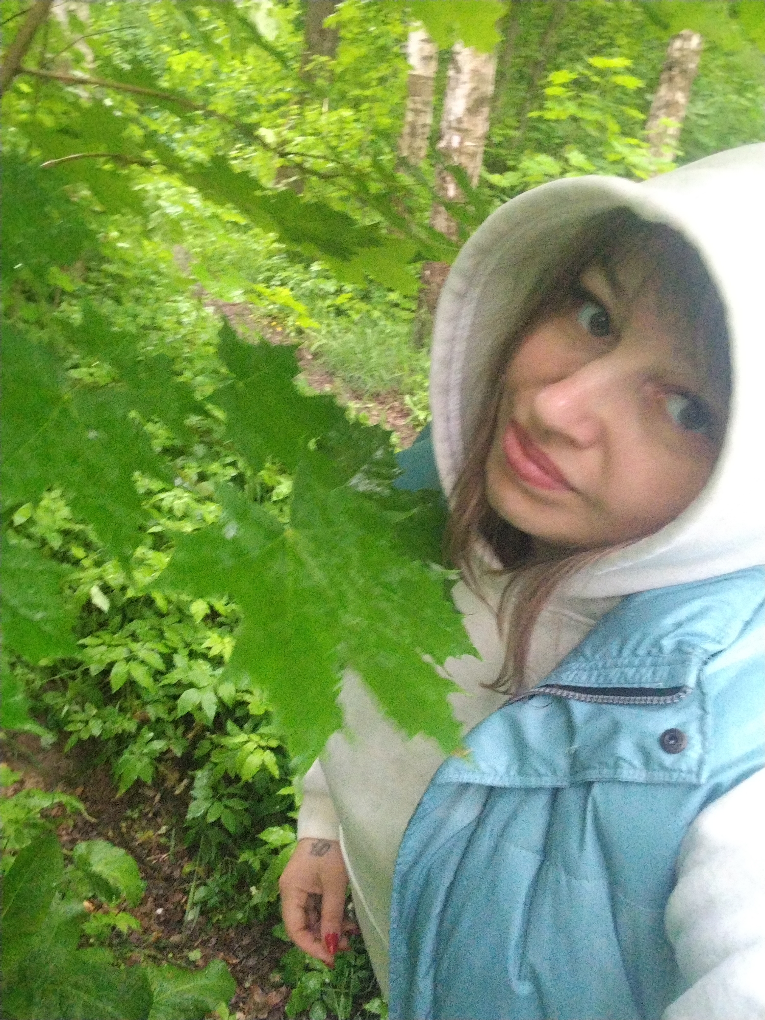 A woman is standing in a forest. She is wearing a white hooded sweatshirt and a light blue vest. The background shows a dense green forest with visible trees. The photo's location is identified as the city of Rusanovo, Russia, based on visual context. The woman is holding a large green leaf in front of her.