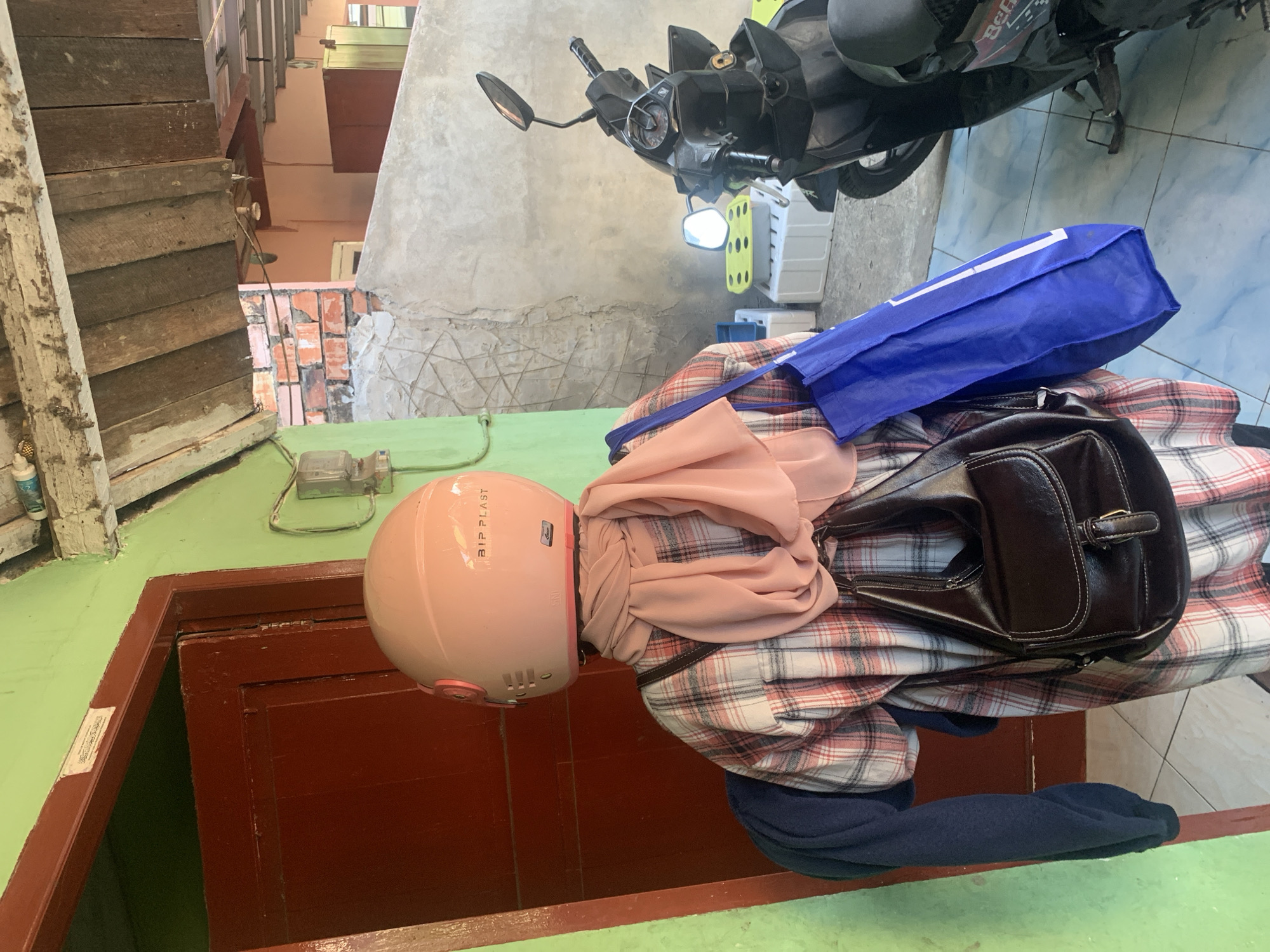 A person wearing a pink helmet, a pink scarf, a plaid shirt, and carrying a dark brown handbag and a blue shopping bag is standing in front of a red door. A navy blue sweater is draped over their left arm. To the right, a black motorcycle is parked. The environment appears to be an outdoor area with a green wall and a wooden roof. The scene is likely in Palembang, Indonesia, suggested by the context of the image.