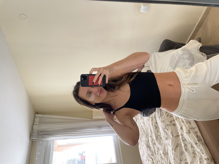 A female individual is depicted taking a mirror selfie in an indoor room within Budva, Montenegro. She stands centered, holding a smartphone with a black case and a red pop-it accessory in her left hand. Her nails are painted black. She wears a black spaghetti-strap crop top, white drawstring sweatpants, and white under-eye patches. Her right hand touches the strap on her right shoulder, where small tattoos are visible. She has long brown hair and a subtle smile. The room features light-colored walls and a tiled floor. To the left, a window with sheer white curtains reveals an exterior building with multi-story, terracotta-colored elements and visible balcony railings, consistent with residential architecture in Budva. A bed with a light-colored duvet patterned with leaves is visible to the left. To the right, a grey upholstered piece of furniture, possibly a chair or sofa, has a light-colored textile with a dark circular graphic draped over it. A white electrical outlet or switch is mounted on the right wall. The scene suggests a private residential or hospitality setting.