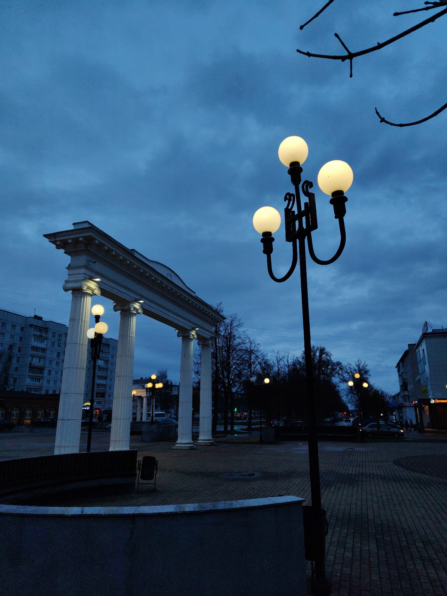 The image depicts a city square at dusk. A neoclassical archway with white columns stands to the left, illuminated by built-in lighting. A black, ornate lamppost with four glowing globe-shaped lights is prominent in the foreground on the right. Buildings are visible in the background, including an apartment building on the left and commercial structures on the right. A park-like area with trees and other lampposts extends into the distance. The ground is paved with brick. The sky is a cloudy, dark blue. There are a bench and a trash can in the center left. Several cars and pedestrians are seen in the distance on the right.