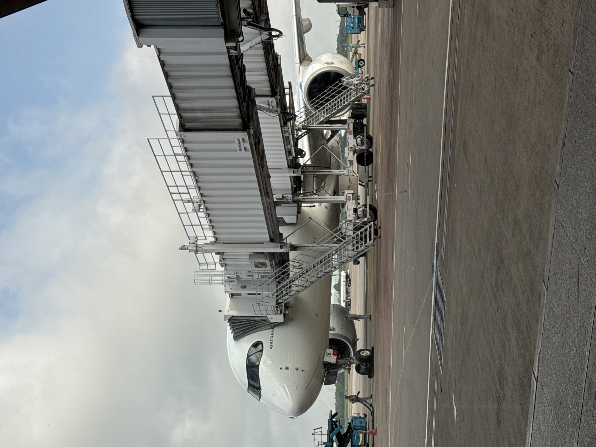 The image shows an Air France airplane parked at an airport gate, connected to a jet bridge. The plane is a large, white commercial jet with its nose facing the viewer. The jet bridge is a long, enclosed walkway with stairs leading up to it. There are several people on the tarmac, some appearing to be ground crew working on the plane. In the background, other airport infrastructure, including vehicles and buildings, can be seen. The sky is partly cloudy. This scene is located at Tan Son Nhat International Airport (SGN) in Ho Chi Minh City, Vietnam, which serves the greater Ho Chi Minh City metropolitan area, including the city of Thuận An.