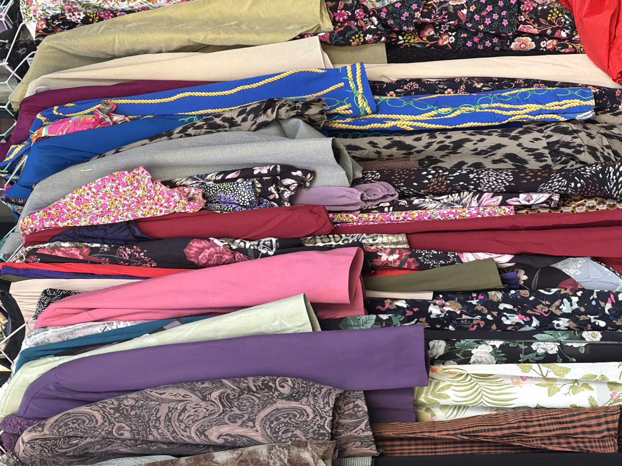 The image shows a stack of various rolled fabrics, likely for sale. The fabrics vary in color, pattern, and texture, including floral prints, paisley, geometric designs, solid colors, and animal prints. The fabrics are stacked on a metal grid or wire shelf. The location is identified as Tbilisi, Georgia, based on the prompt. The scene depicts a textile shop or market setting with no apparent individuals visible in the frame. The fabrics are the primary focus, presented in a disorganized and layered arrangement.