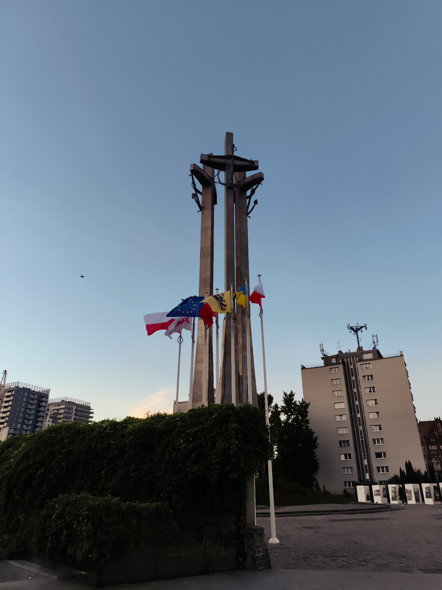 This is a low-angle, wide shot of the Monument to the Fallen Shipyard Workers of 1970 in Gdańsk, Poland. The monument, a tall structure with three large crosses, stands against a clear, pale blue sky. Several flags are visible in front of the monument, including the flag of the European Union, the Polish flag, and two other flags, one yellow and one that appears to be red and white. To the right of the monument is a modern, light-colored apartment building with many windows. In the foreground, there is a cobblestone plaza and a dense, dark green hedge. In the distance, to the left, are other modern buildings, one of which appears to be under construction. A single bird is visible flying in the sky to the left of the monument. The lighting suggests it is either early morning or late evening.