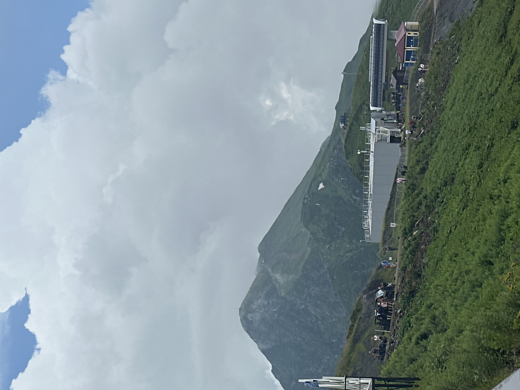 In the city of Estosadok, Russia, a mountainous landscape features a prominent green peak under a sky filled with white clouds. A cable car station with a long, white carriage is visible on the right side of the frame, situated on a grassy slope. Adjacent to the station are a few small buildings, one with a red roof and another with a light-colored facade. A group of people are gathered near the station and on the slope, some standing around vehicles, while others appear to be walking or observing the scenery. The slope is covered in lush green vegetation, with some rocky outcrops interspersed. In the distance, the mountain exhibits green slopes and signs of a path or road winding up its side.
